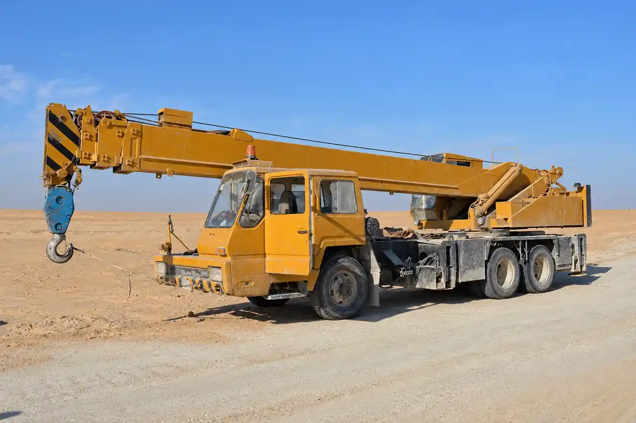 Yellow heavy-duty mobile crane with an extended boom and hanging hook parked on a dusty desert road.
