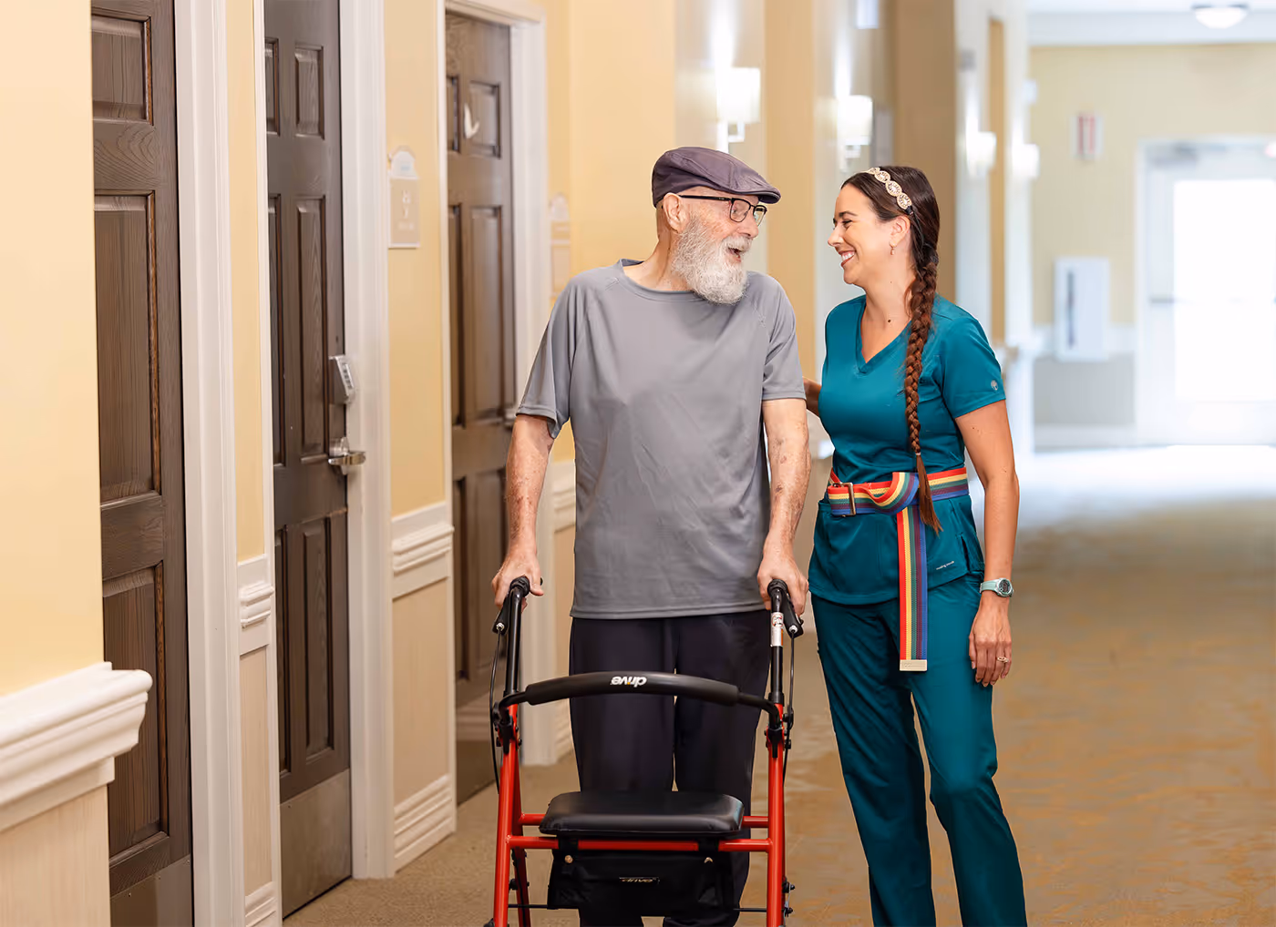 Elderly man with a walker talks and smiles with a caregiver in a hallway.