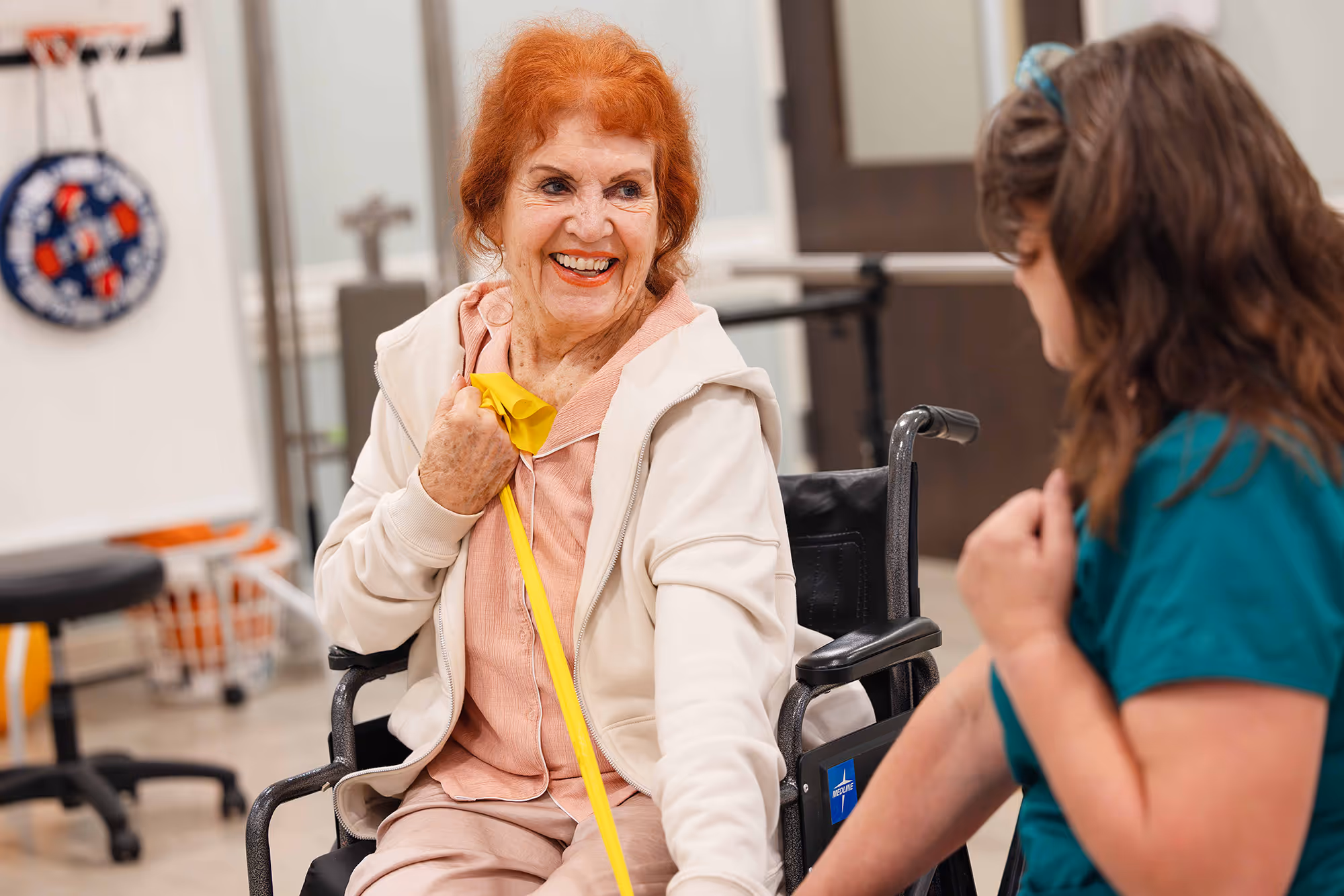 Older woman in a wheelchair using a yellow resistance band with assistance from a caregiver.