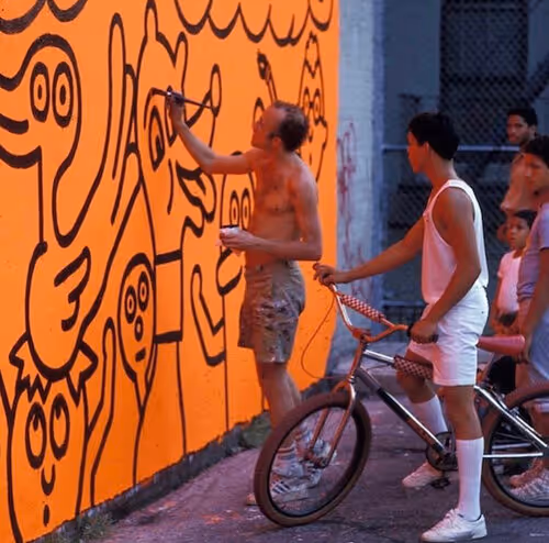 Artist painting cartoon-style mural on bright orange wall while person on bicycle watches