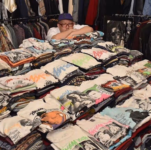 Person with glasses and purple cap behind large collection of folded graphic t-shirts