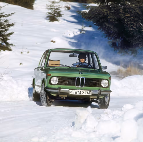 Vintage green BMW 2002 driving through snowy forest road with winter landscape backdrop