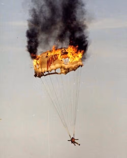 Parachutist descending beneath a burning canopy with billowing black smoke against a pale sky