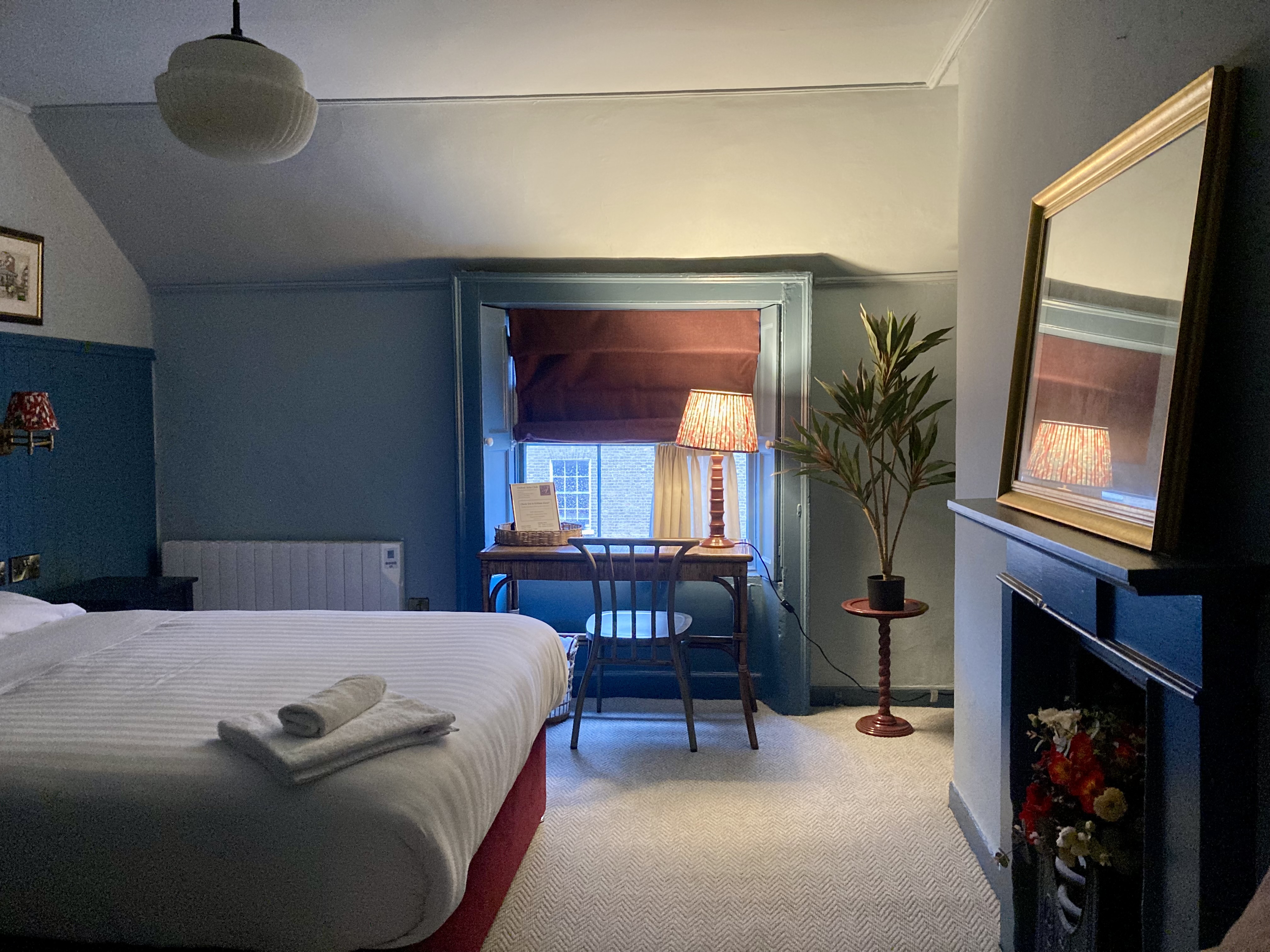 Bedroom interior featuring a neatly made bed with a yellow throw, a decorative fireplace, artwork above the mantlepiece, and a red armchair with a colourful cushion.