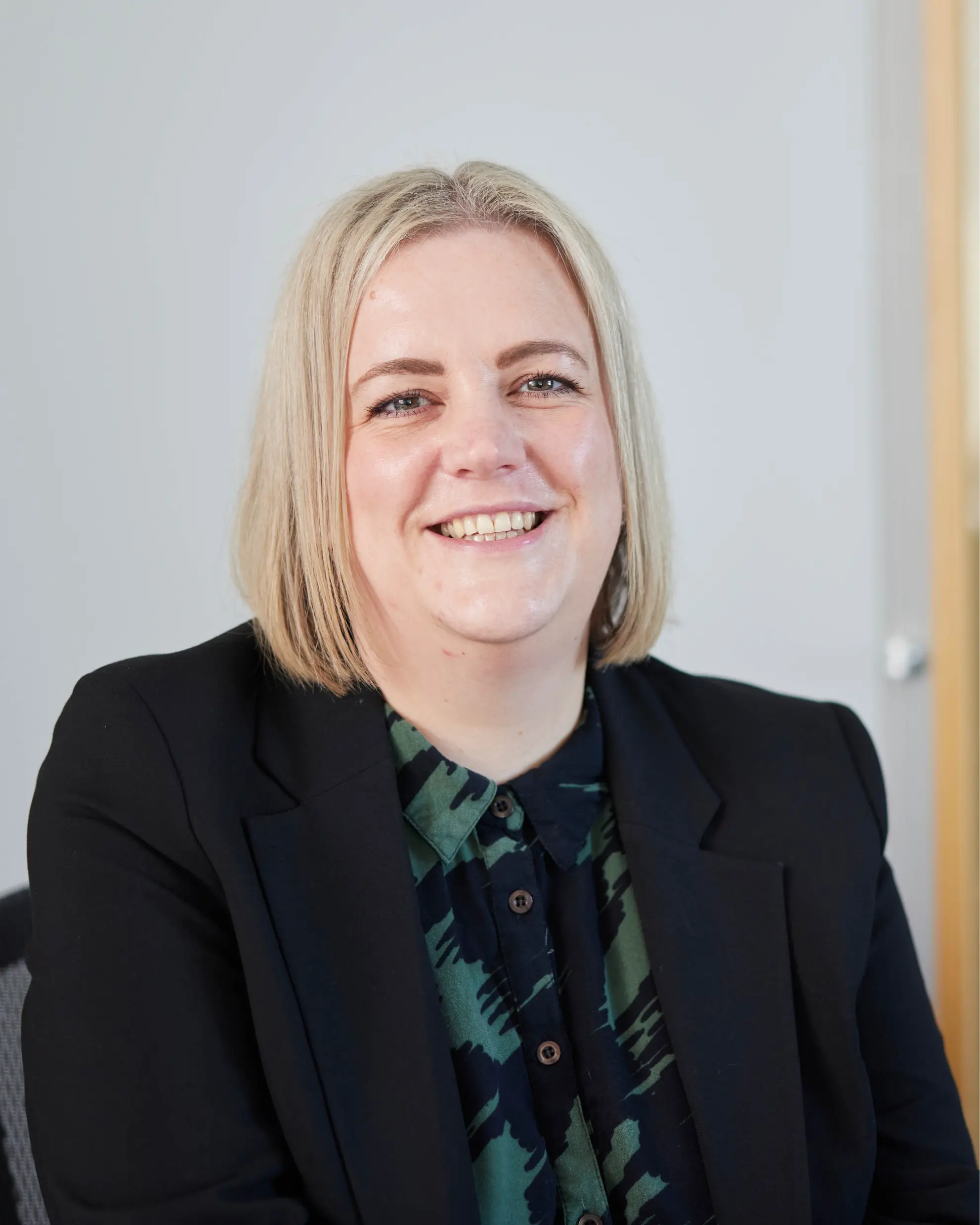 Smiling woman with shoulder-length blonde hair wearing a black blazer and patterned green and black shirt.