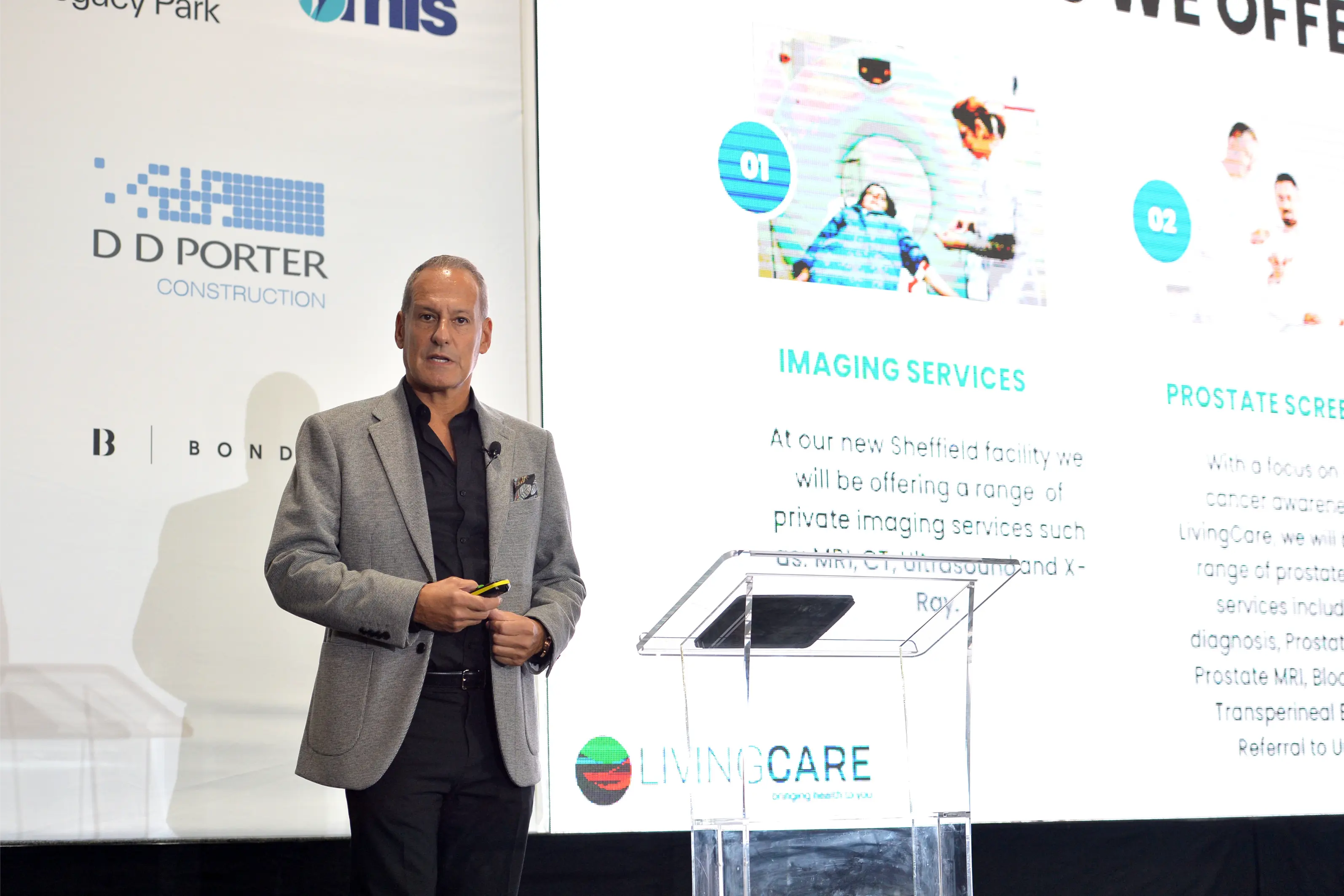 Man in gray blazer and black shirt giving a presentation next to a screen showing imaging and prostate screening services.