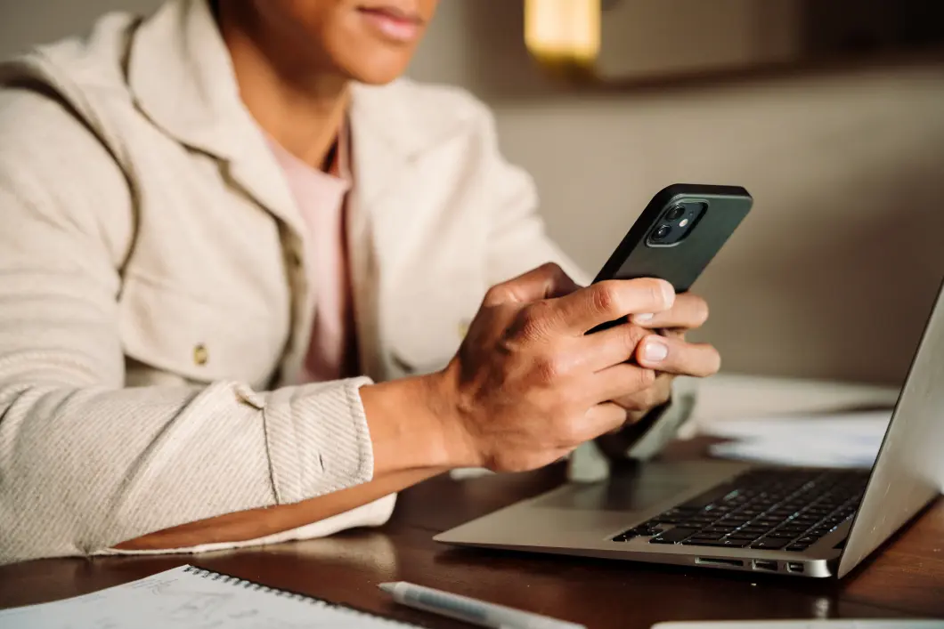 Person holding a smartphone near an open laptop on a wooden desk with papers and a pen.