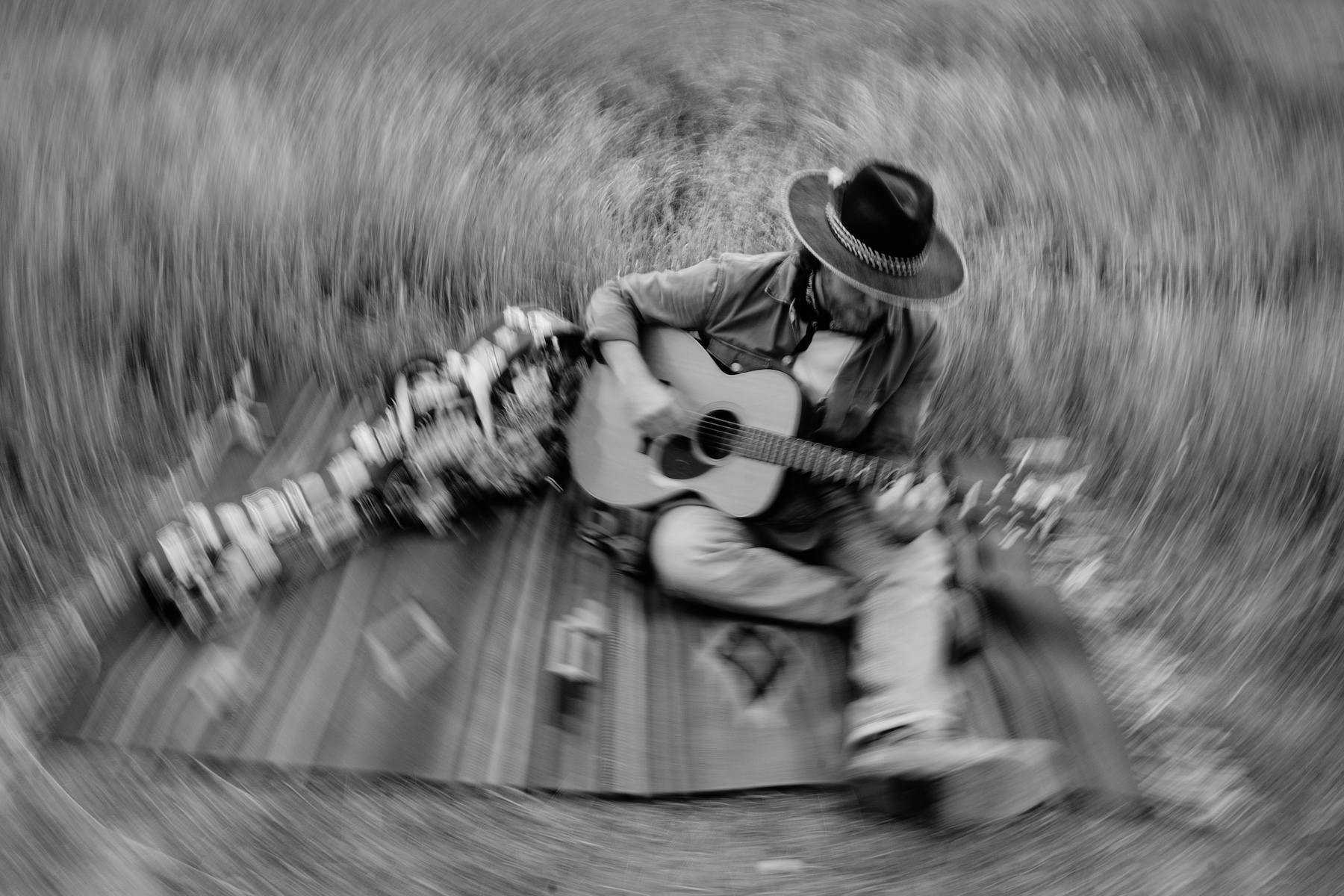 Black and white photo of a man wearing a hat playing an acoustic guitar while sitting on a blanket outdoors with a blurred, swirling effect.