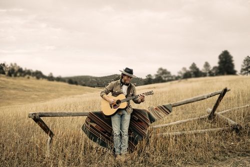Man wearing a hat playing an acoustic guitar while leaning against a rustic wooden fence in a dry grassy field.