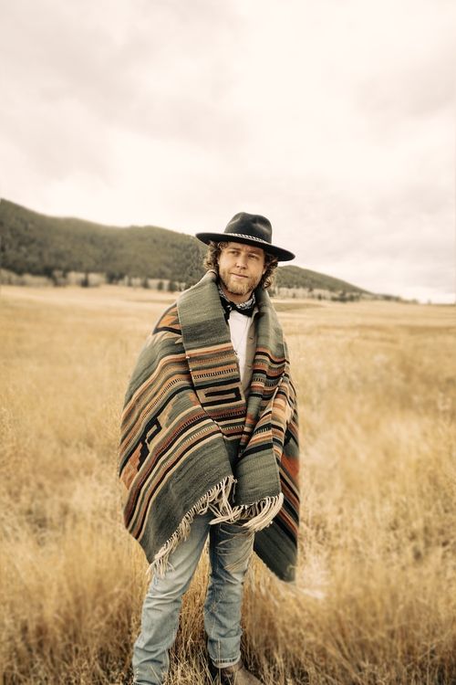Man in a black hat wrapped in a patterned blanket standing in a dry grassy field with hills in the background.
