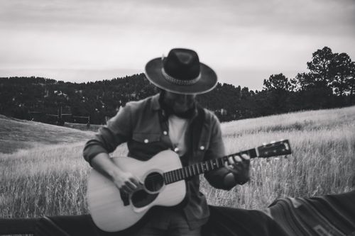 Person wearing a wide-brimmed hat playing acoustic guitar outdoors in a grassy field with trees and hills in the background.