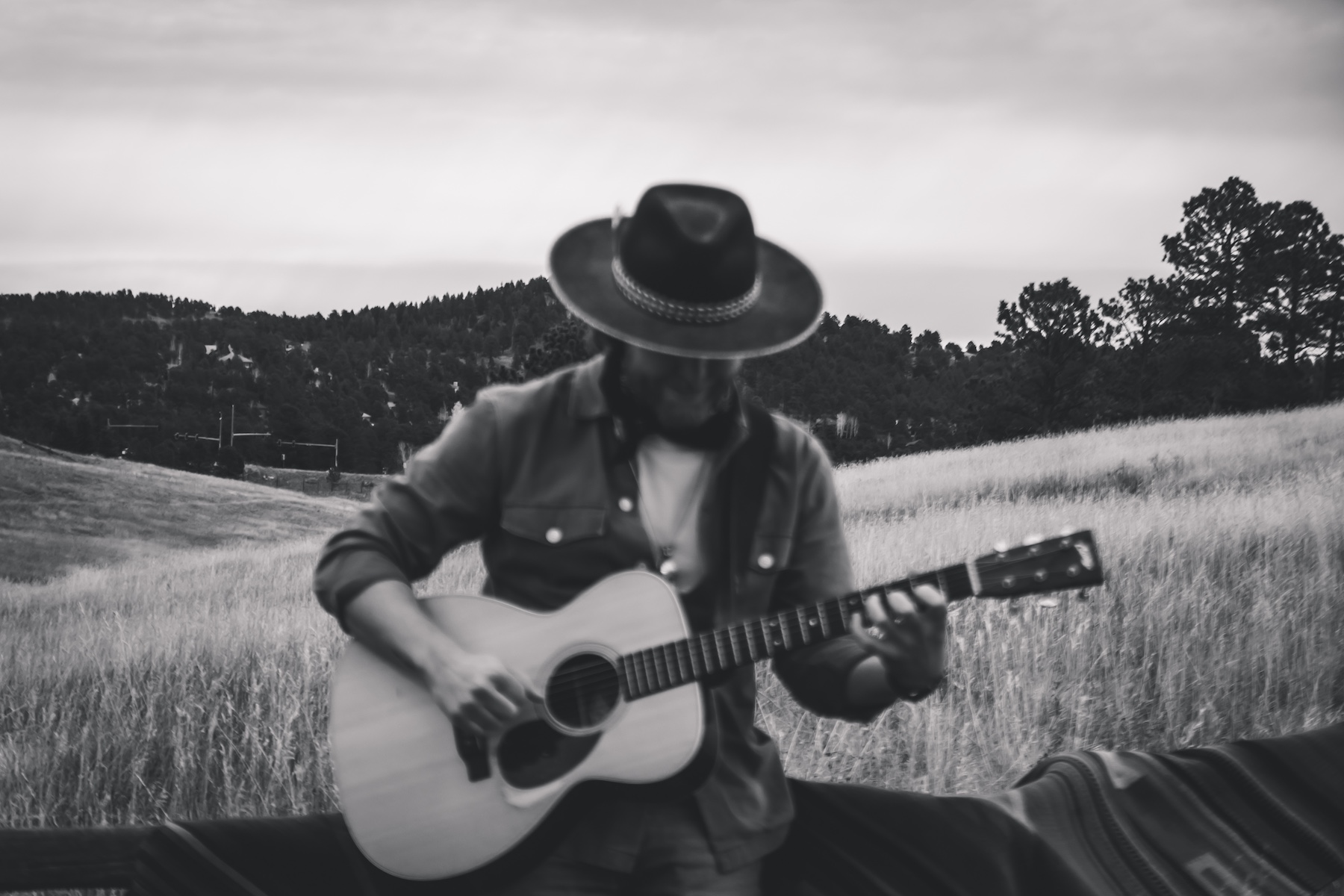 Person wearing a wide-brimmed hat playing acoustic guitar outdoors in a grassy field with trees and hills in the background.