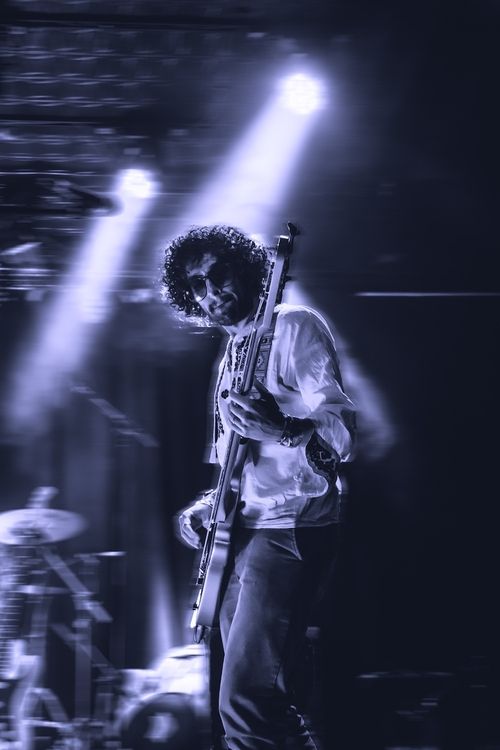 Musician with curly hair and sunglasses playing electric guitar on stage under bright spotlights.