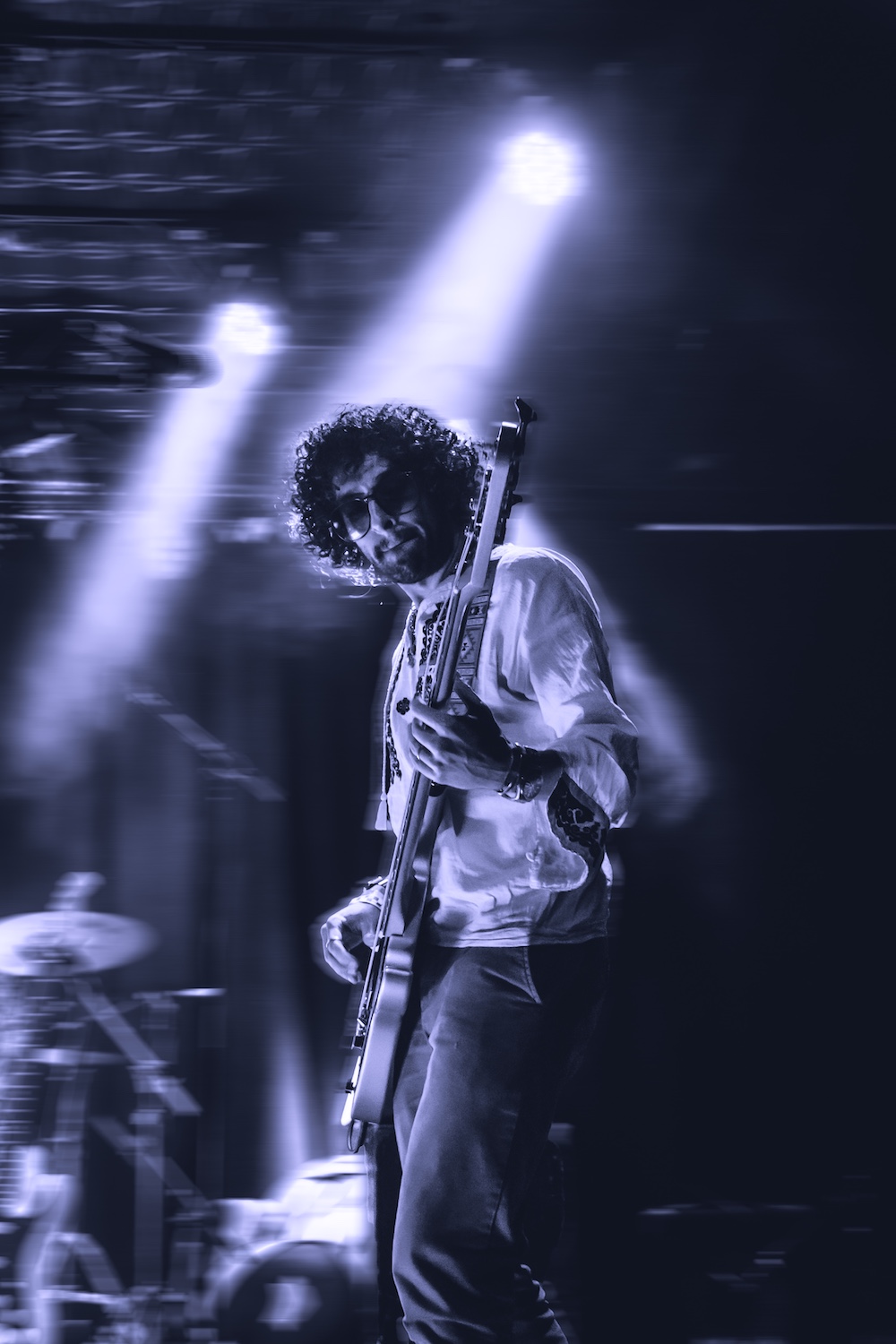 Musician with curly hair and sunglasses playing electric guitar on stage under bright spotlights.