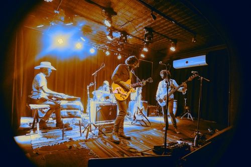 Band performing live on stage with guitar, bass, drums, and keyboard under warm stage lights.