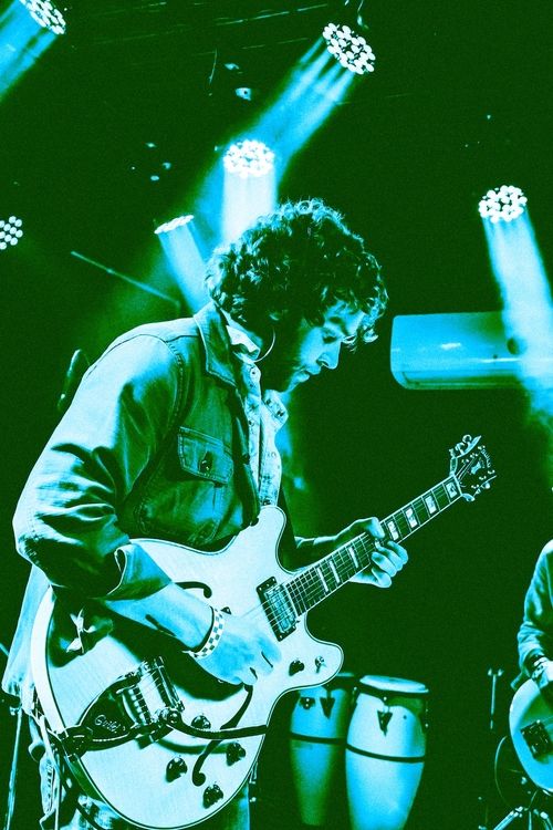 Male guitarist playing an electric guitar on stage under blue and green lighting.