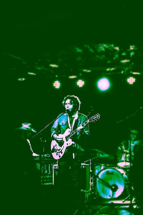 Musician with curly hair playing guitar and singing on stage with band members and drum set under bright stage lights.