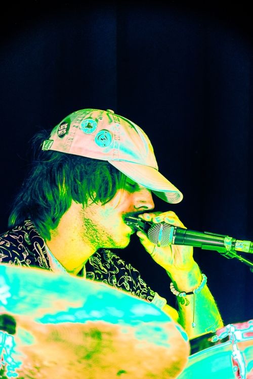 Musician wearing a cap with pins playing a harmonica into a microphone against a dark background.
