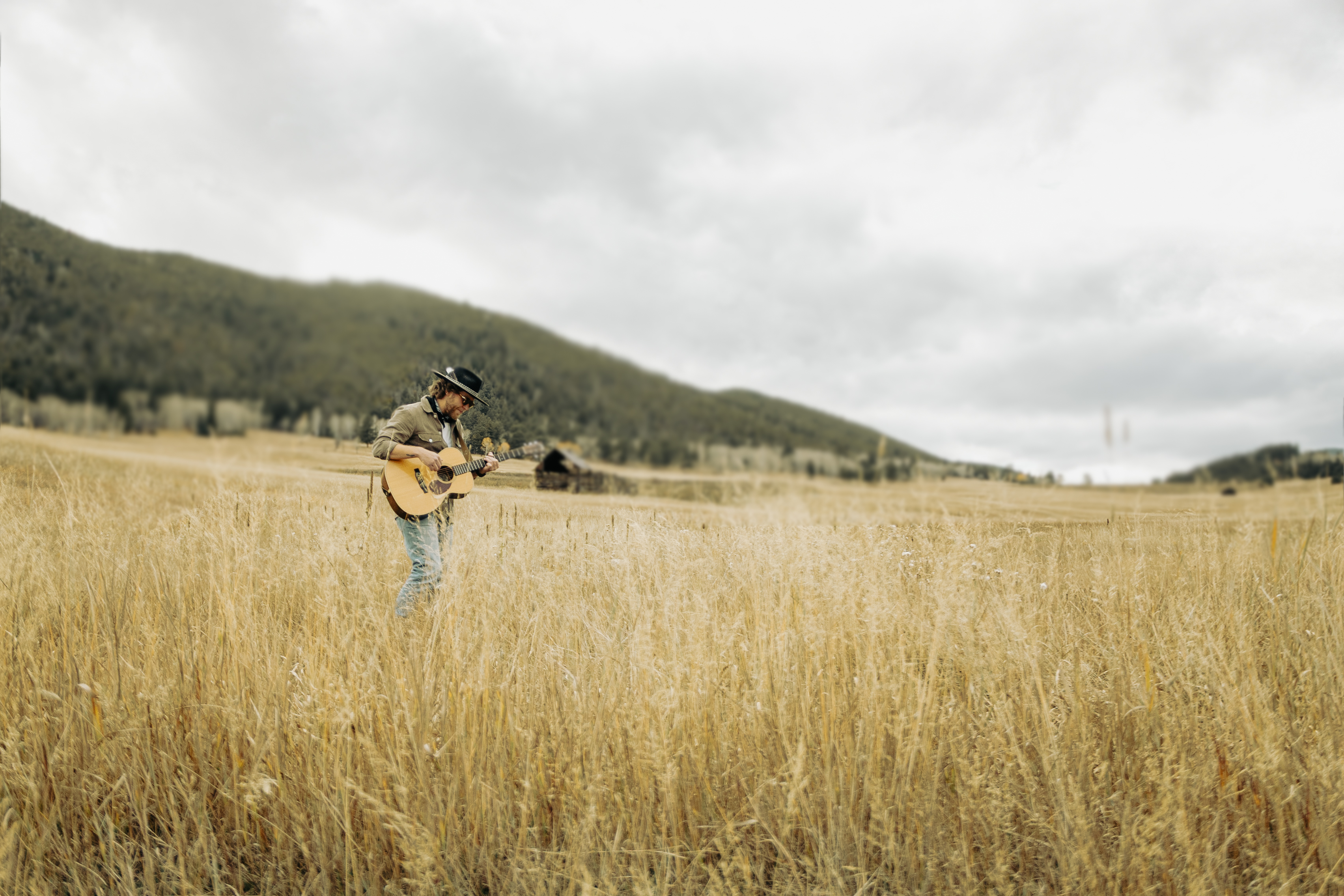 Man wearing a hat playing an acoustic guitar standing in a tall dry grass field with hills and cloudy sky in the background.