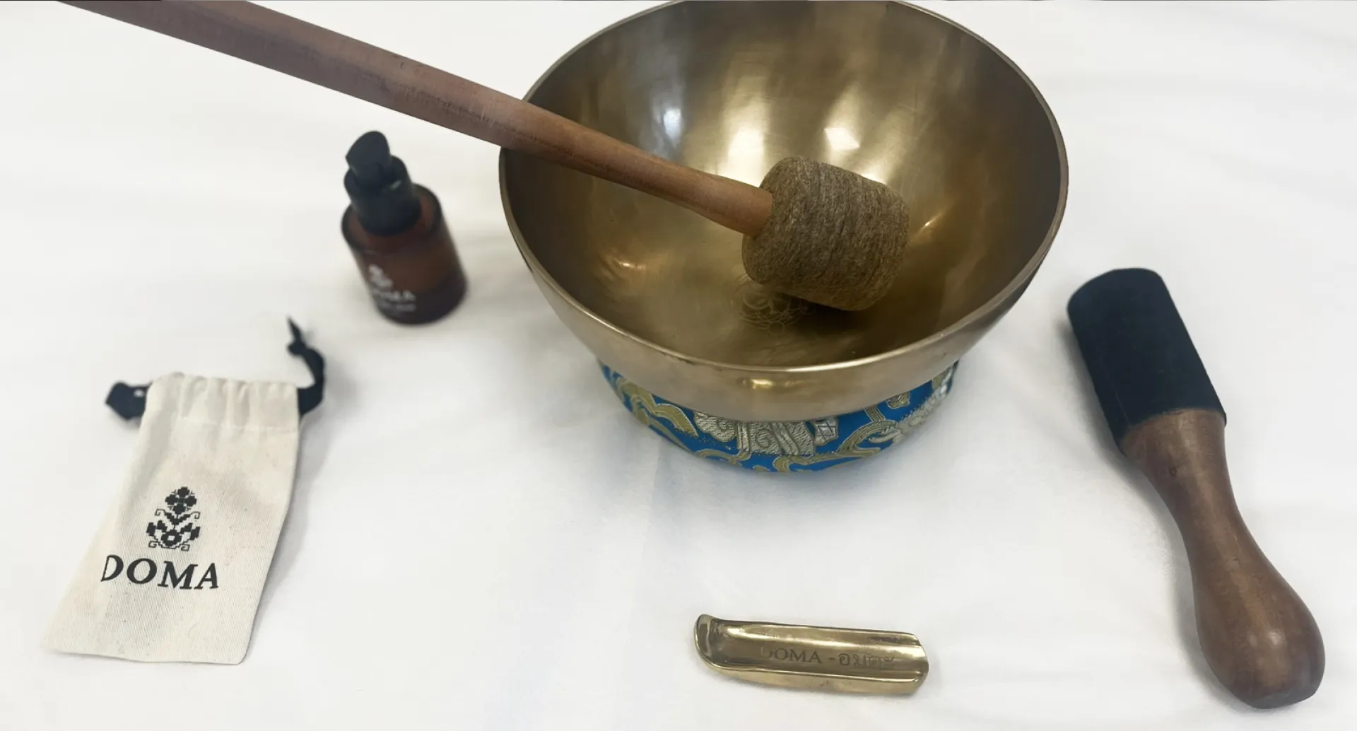 Brass singing bowl with striker, wooden mallet, small pouch, and brown dropper bottle on white surface.