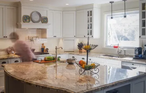 Full view of white kitchen cabinets and large marble island, blurred homeowner moving around kitchen