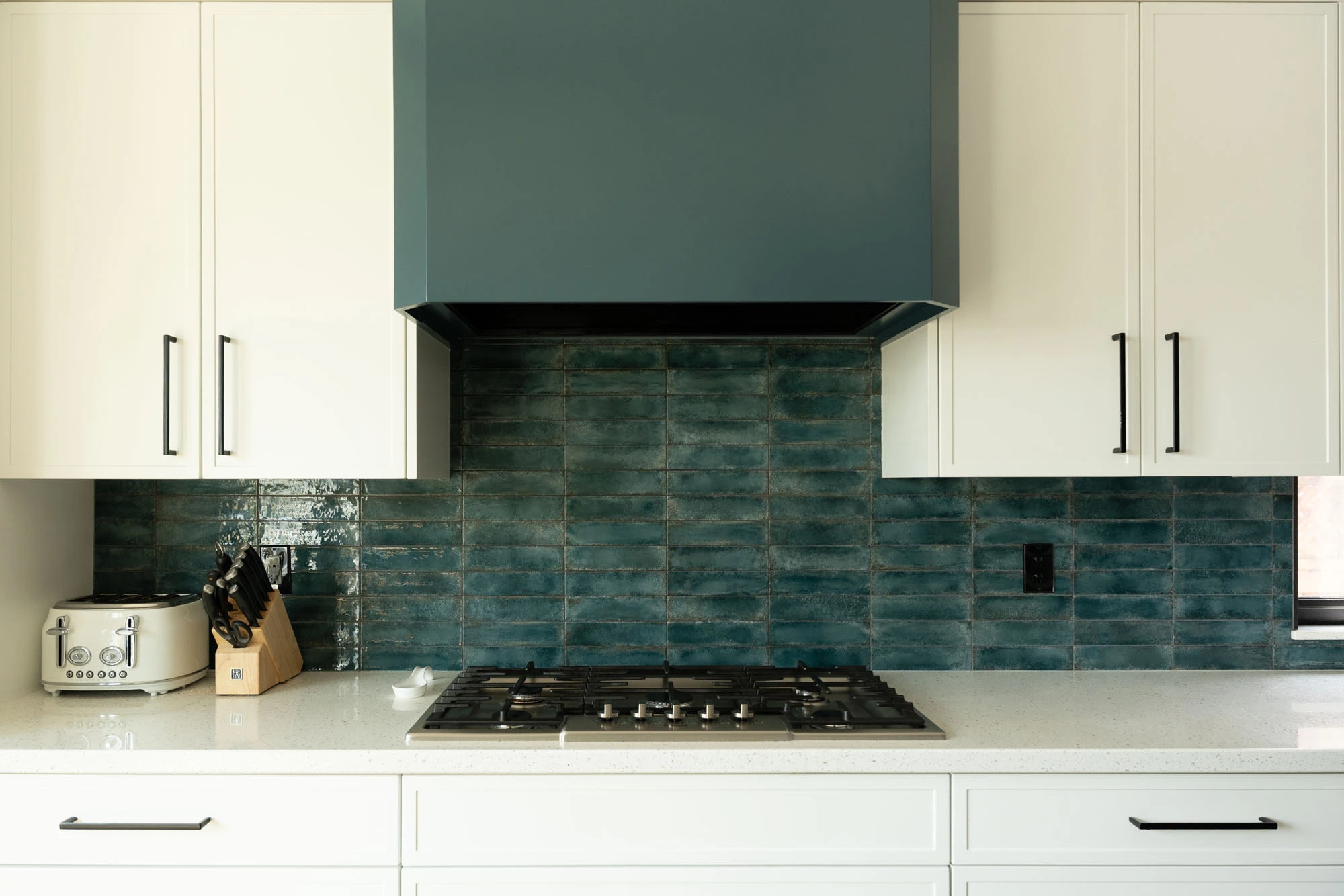 Detail shot of wall of kitchen cabinetry featuring white cabinets, a blue range hood, and matching blue tile backsplash