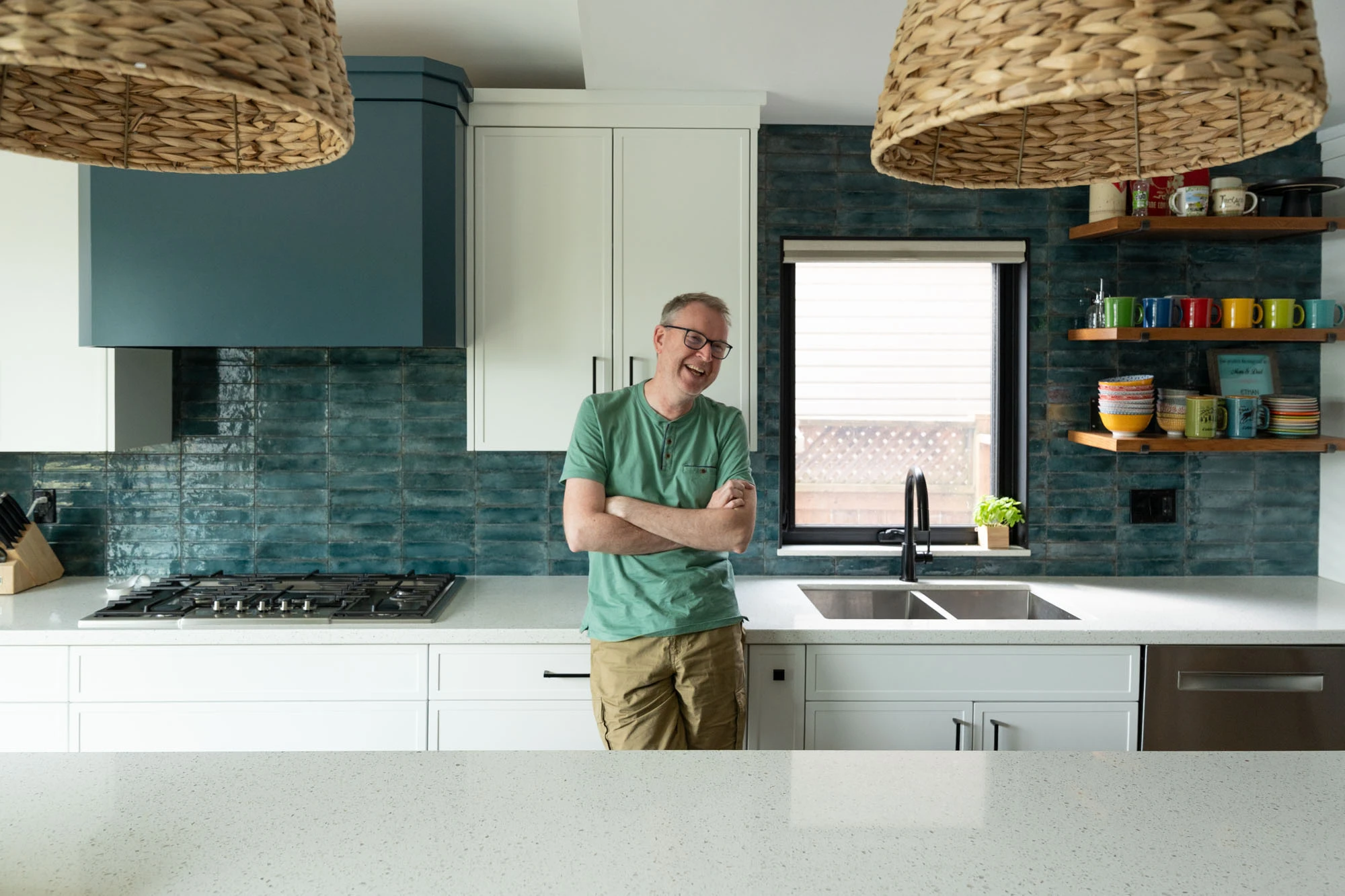 Smiling homeowner leaning against kitchen countertop with arms crossed. Stovetop and kitchen sink in background