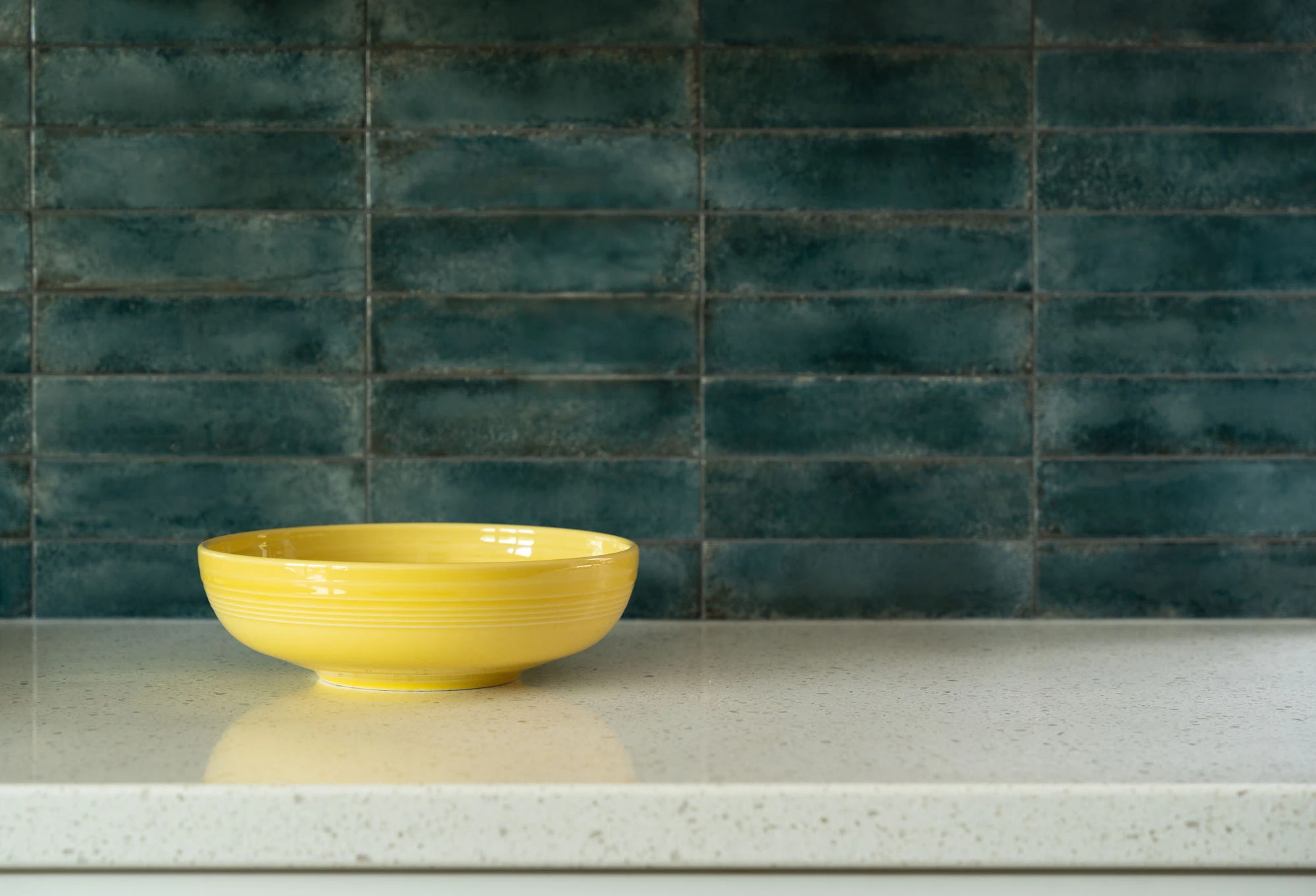 Bright yellow ceramic bowl sitting on white kitchen countertop, blue tile backsplash in the background