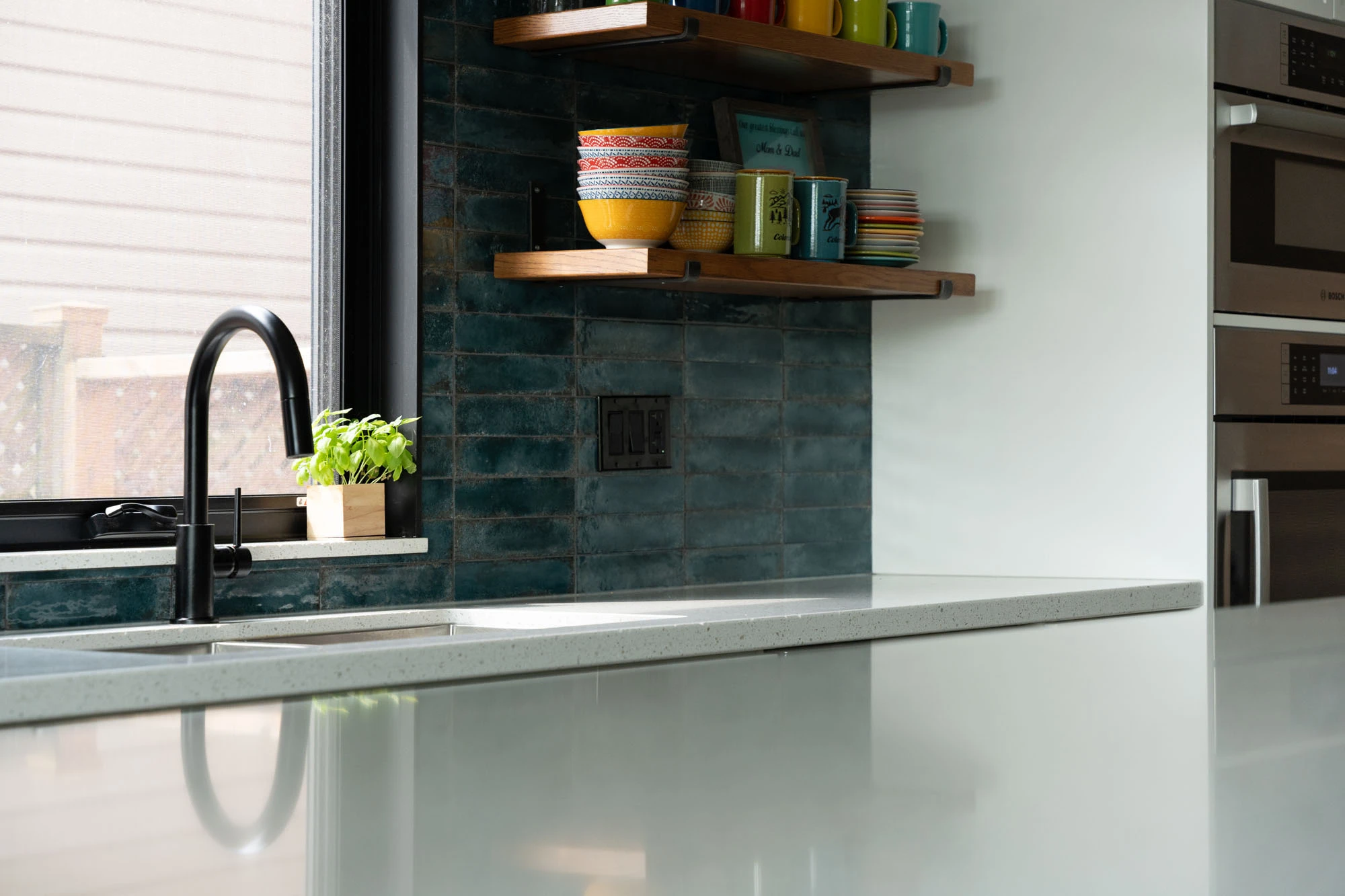 White kitchen countertops in foreground with kitchen sink and colourful ceramic dishes on shelving in background