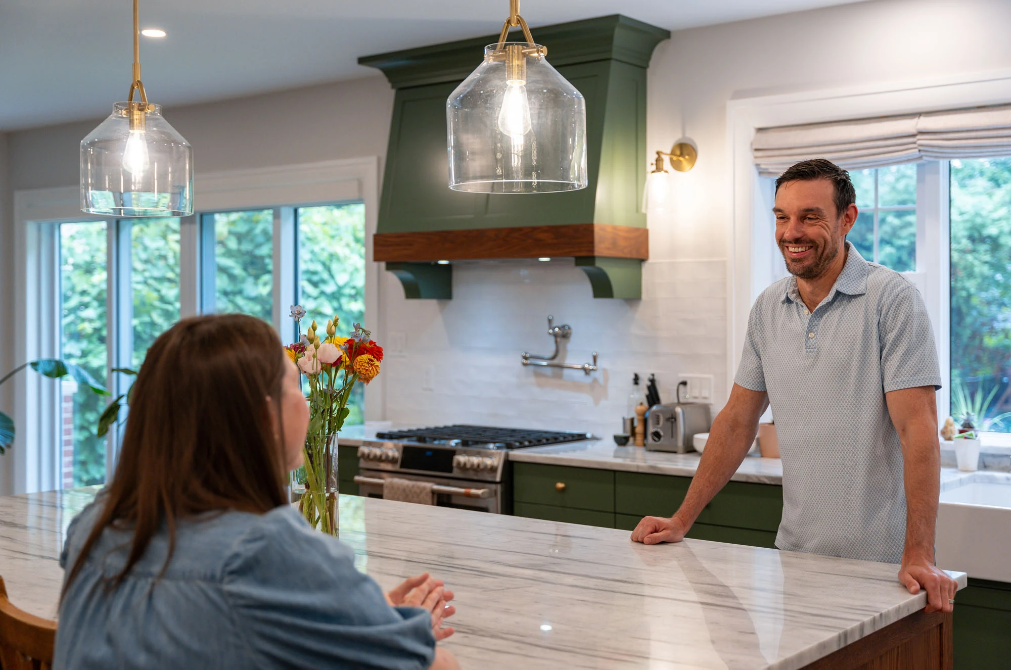 Homeowners at kitchen island, one stands and leans on countertop while other sits on wooden stool.