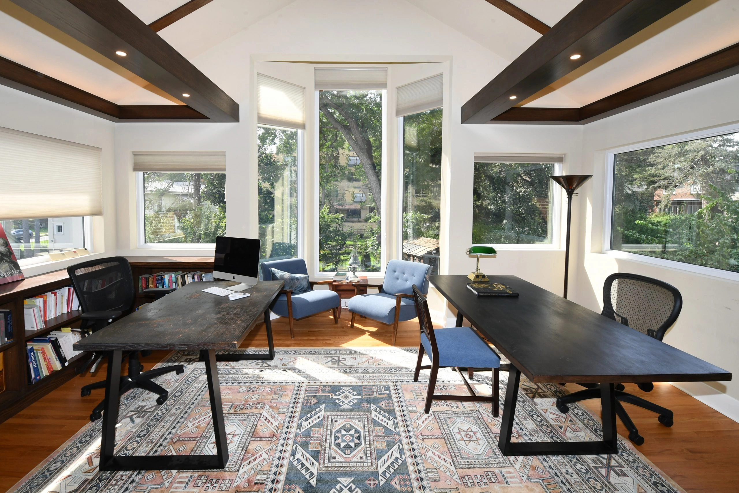 An inviting library with large windows casting light onto a desk and chair with a wall of books behind it