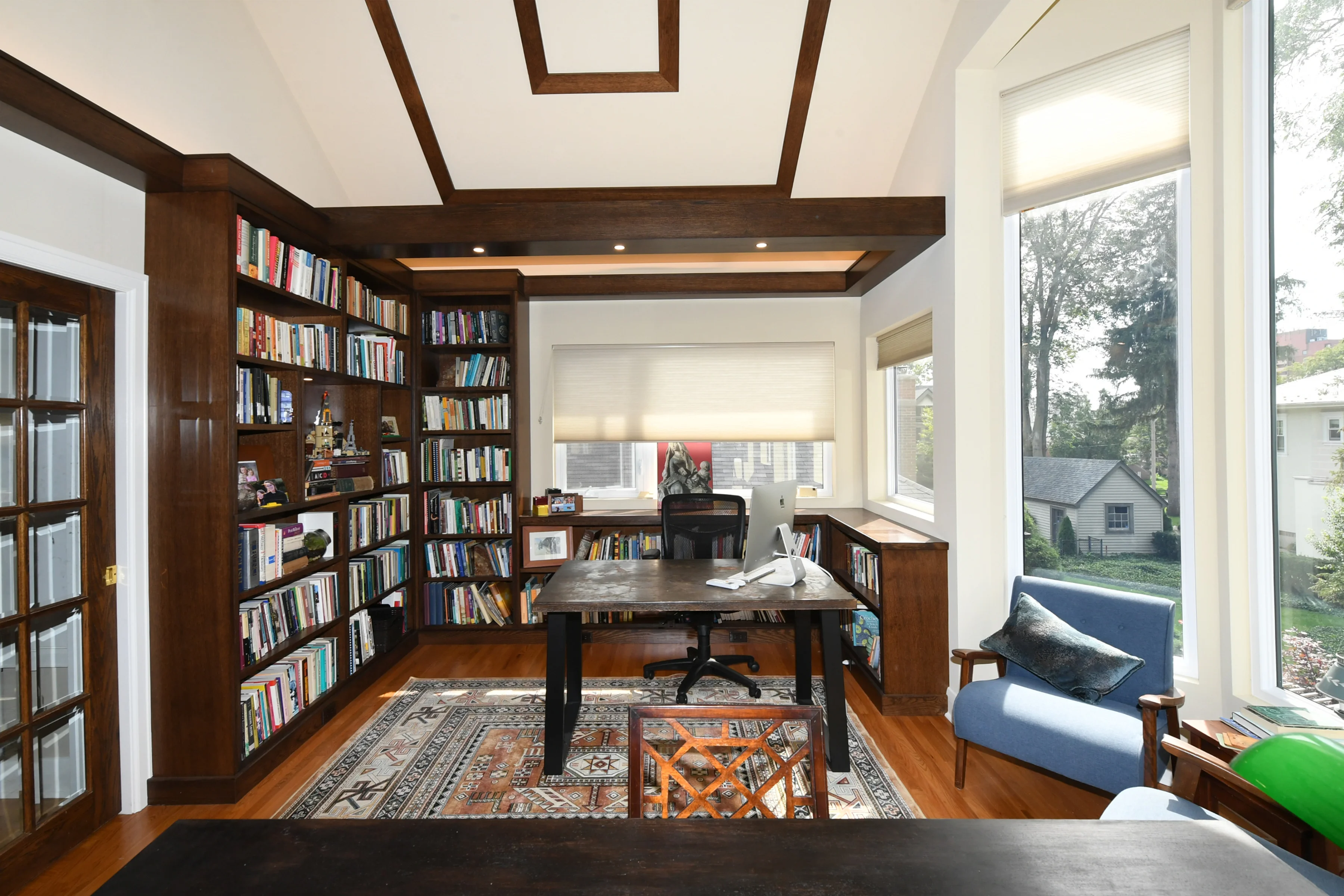 An inviting Library with large windows letting in sun, with a desk and chairs for sitting in