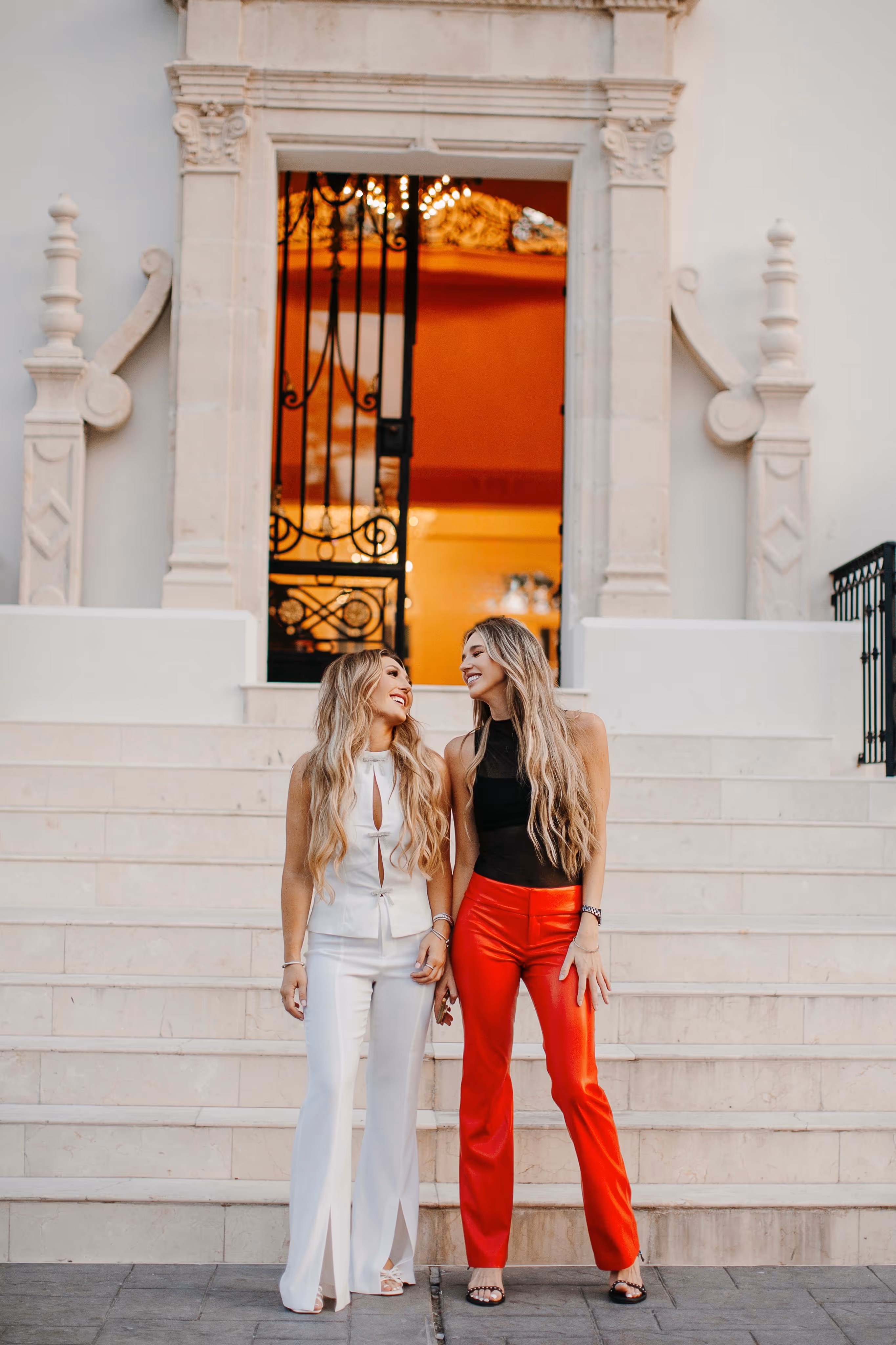 Two women with long blonde hair smiling at each other, standing on stairs in front of a building, one wearing white outfit and the other red pants and a black top. Hope and Marxli Social Habit Marketing Cofounders of digital marketing agency