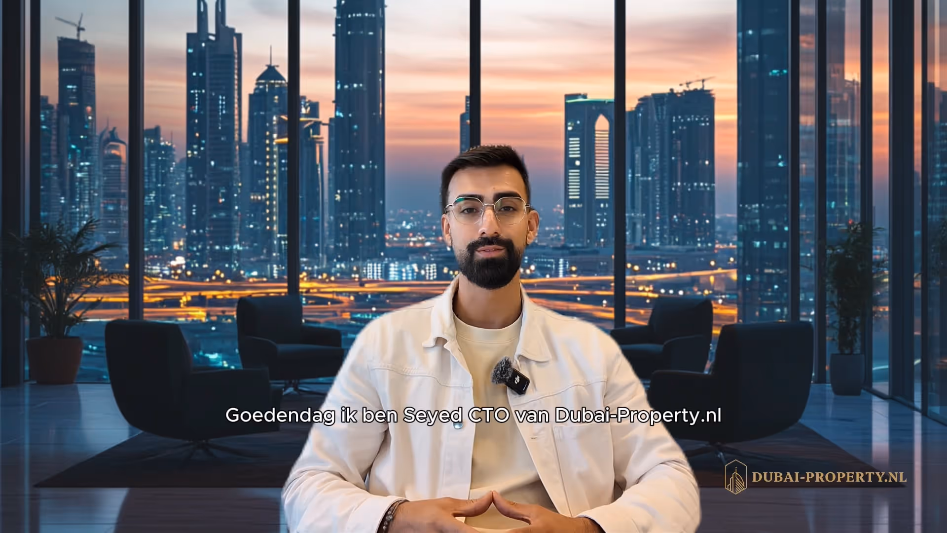 Man met bril en baard zit in een moderne kantoorruimte met uitzicht op de verlichte skyline van Dubai bij zonsondergang.