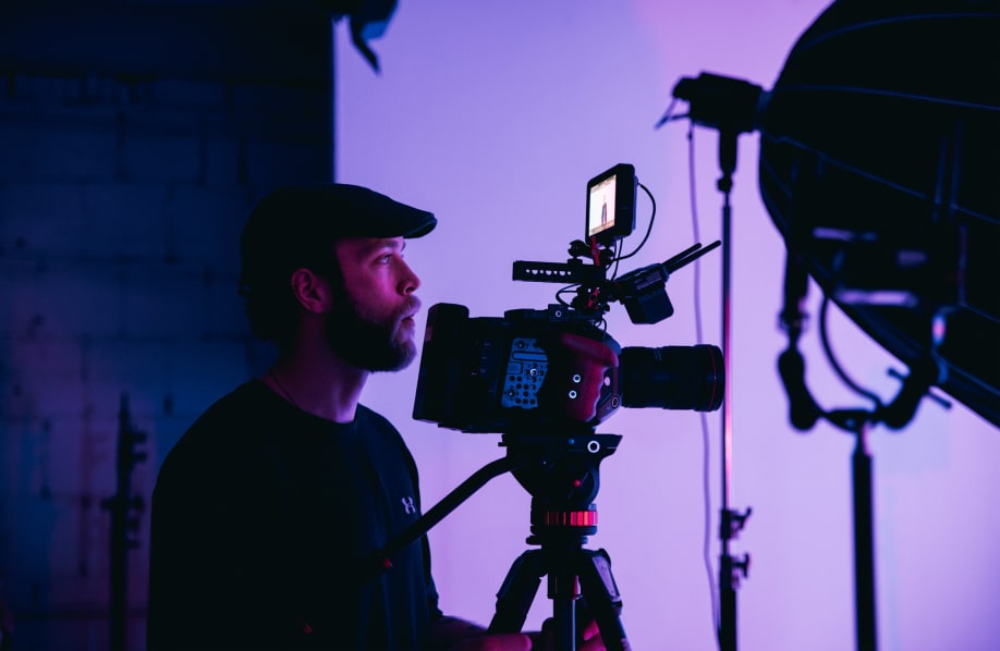 Man with a beard wearing a cap operating a professional video camera on a tripod in a studio with purple lighting.