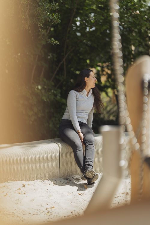 A woman sitting on the 3D-printed sandbox bench, demonstrating its functionality and seamless integration into the natural playground environment.
