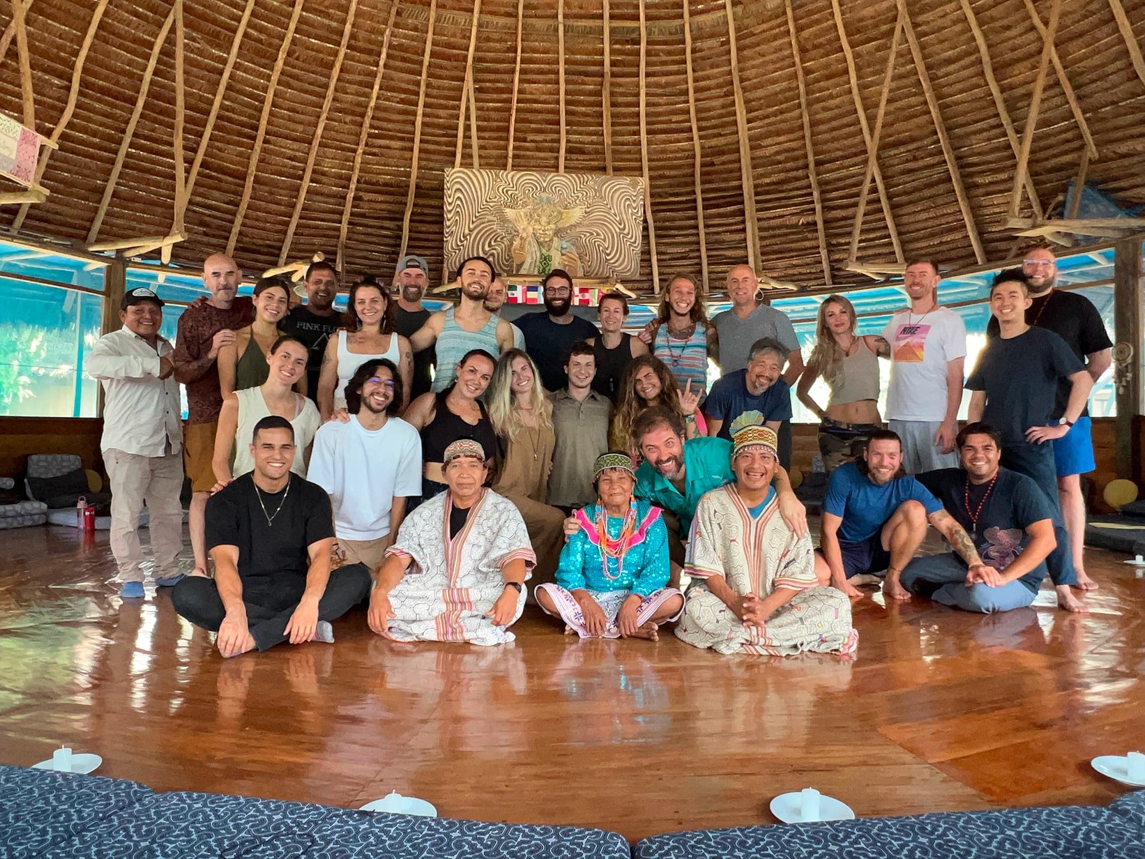 A group of people sitting in a circle inside a ceremonial hut, with a shaman in the center.
