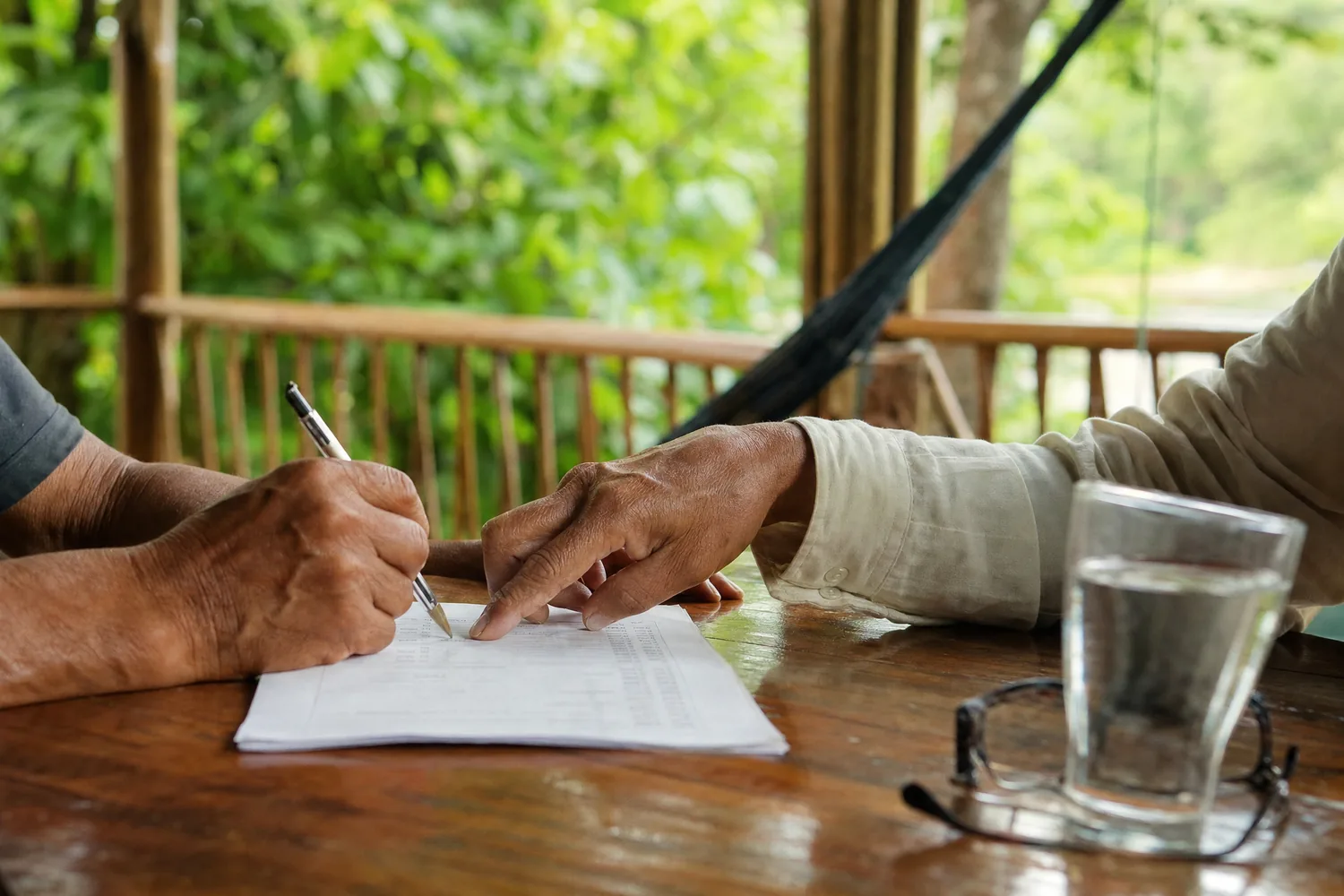 Two people's hands at a wooden table on a rainforest veranda, one completing a paper health questionnaire while the other points to a line