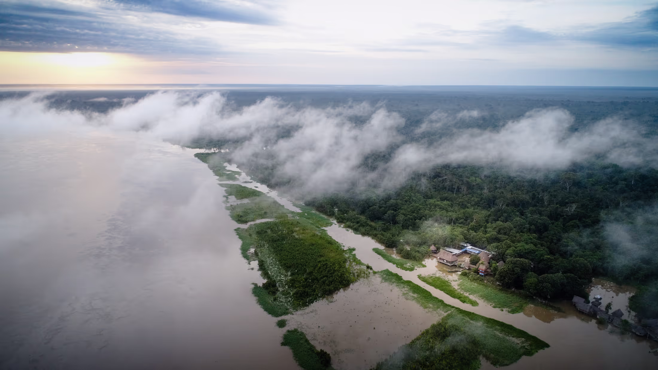 Aerial view of Arkana's Amazon retreat center beside the river, with mist over the rainforest at sunrise