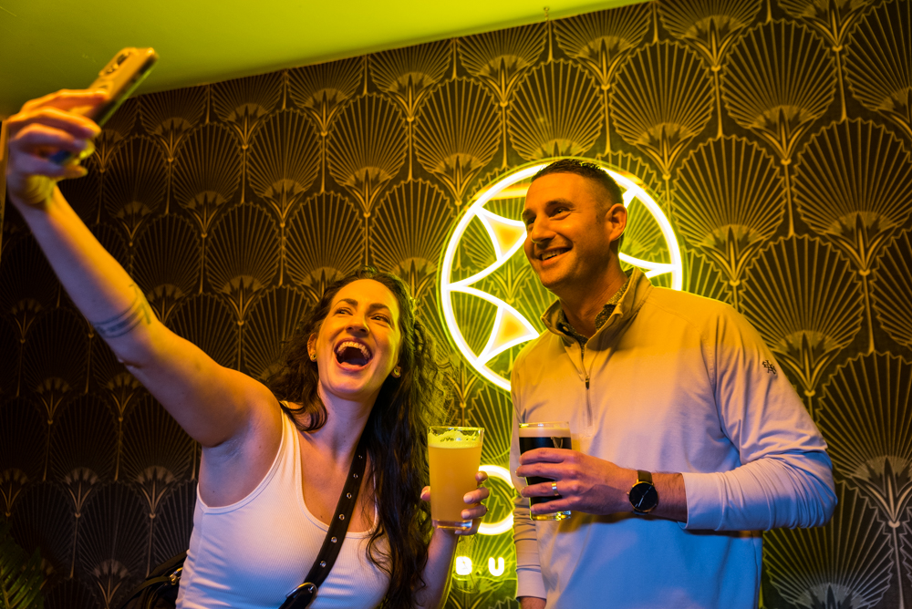 Two friends taking a selfie with drinks in front of The Caz neon sign