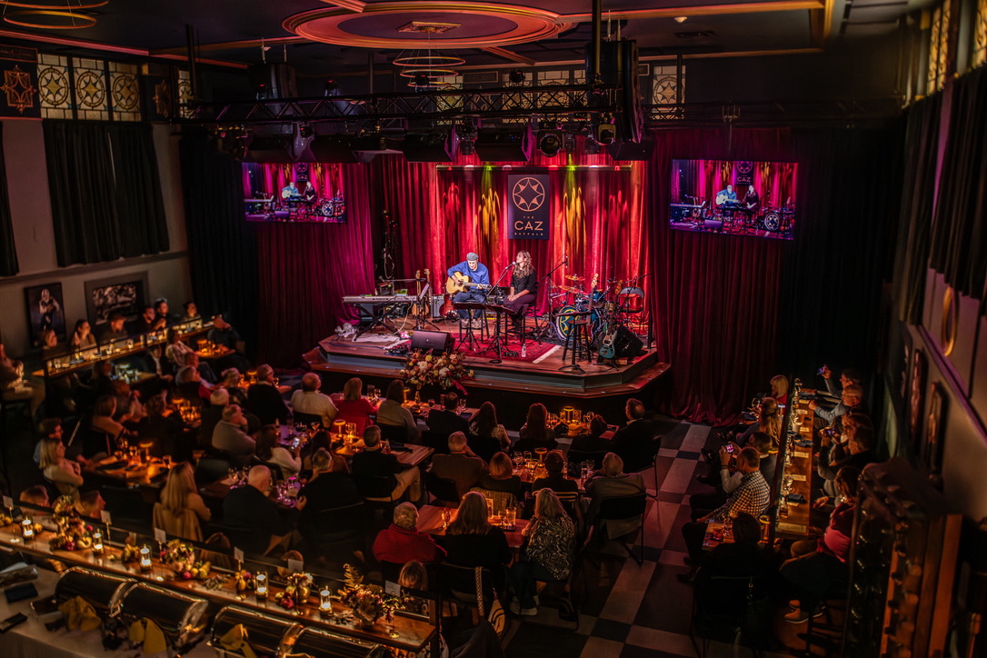 A band performing on stage to a full dining room at The Caz, seen from the mezzanine
