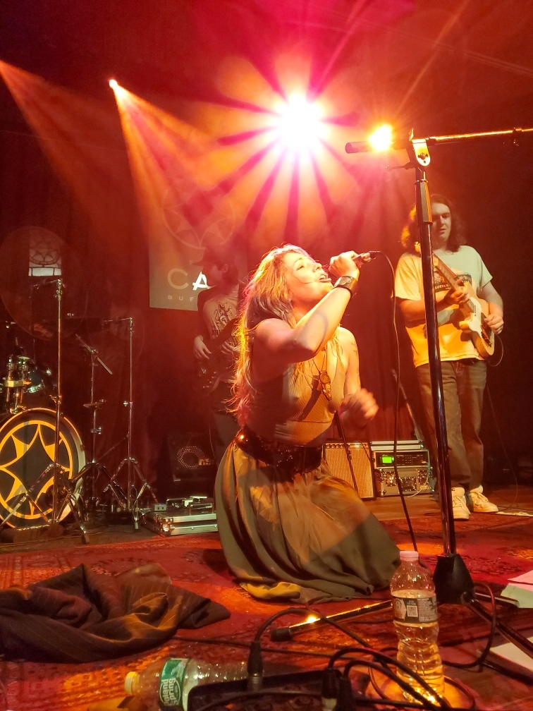 A singer kneeling at the front of the stage under warm lights at The Caz