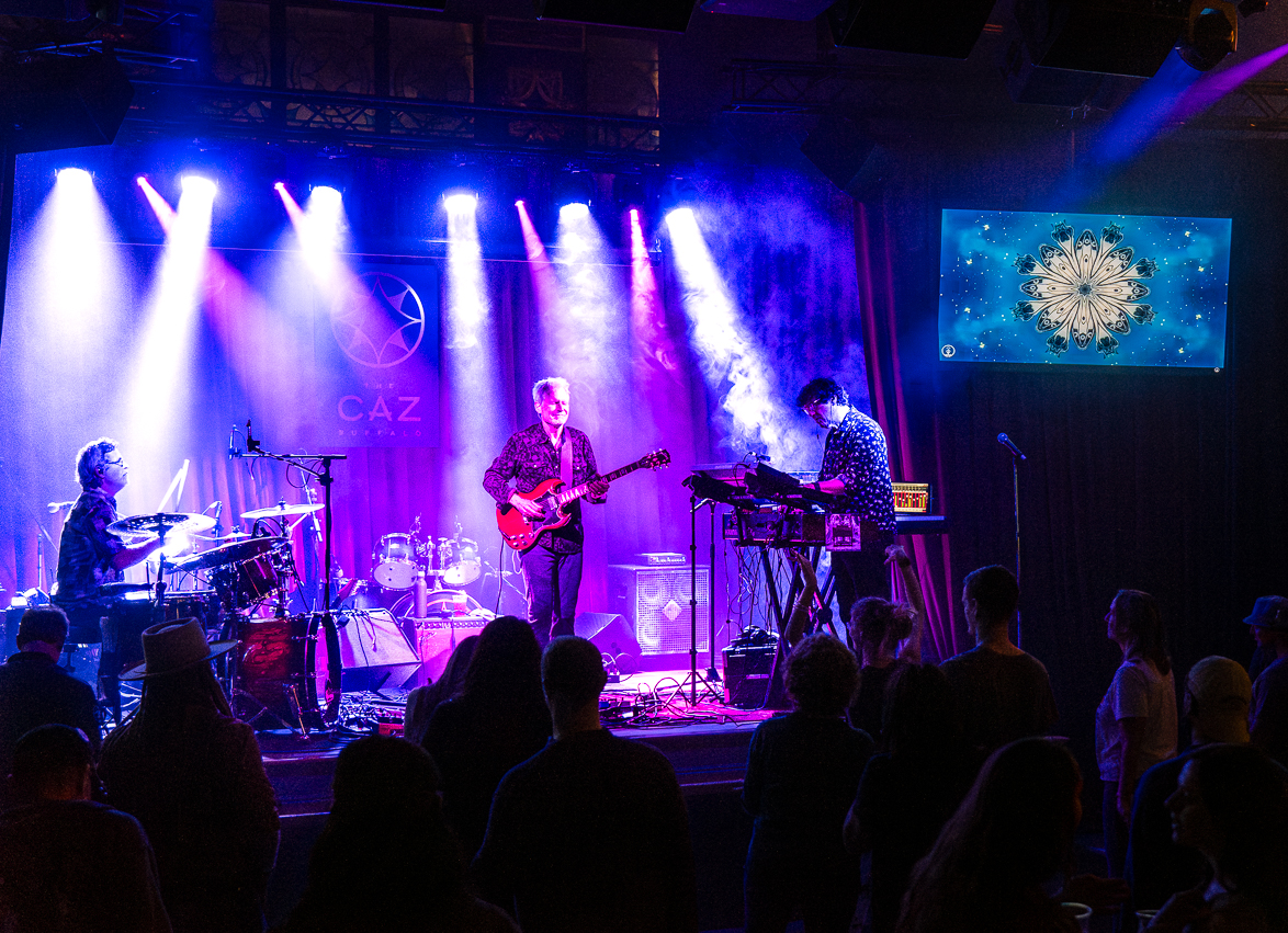 A band performing under blue stage lights to a standing crowd at The Caz