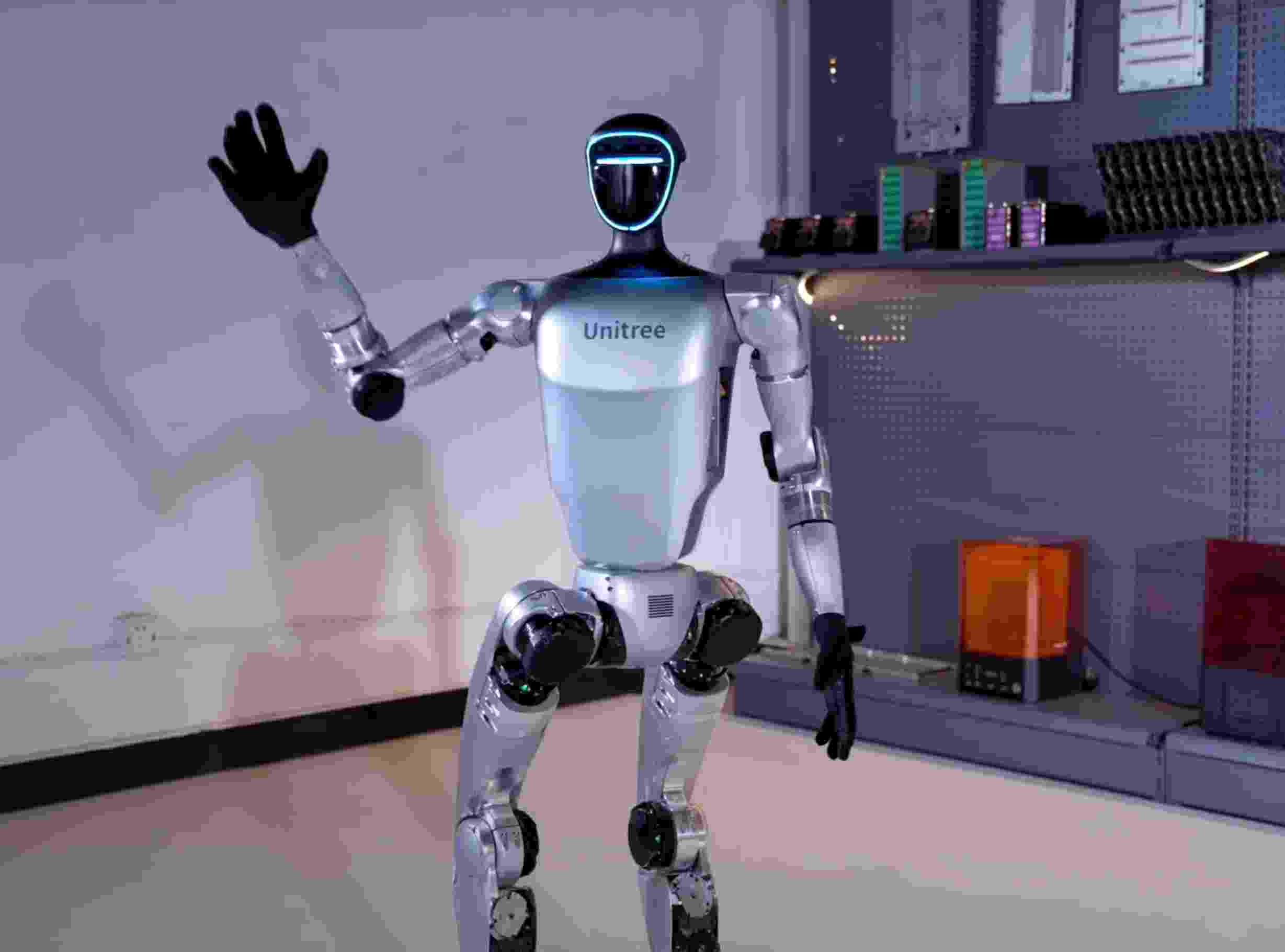 Unitree G1 humanoid robot in a research laboratory environment, standing with one arm raised in a waving gesture. The robot is positioned in front of laboratory equipment including shelving units with electronic components and testing equipment. The 'Unitree' branding is visible on the robot's silver torso. This image demonstrates the G1's deployment in real research and development settings where universities and corporations conduct robotics experiments.