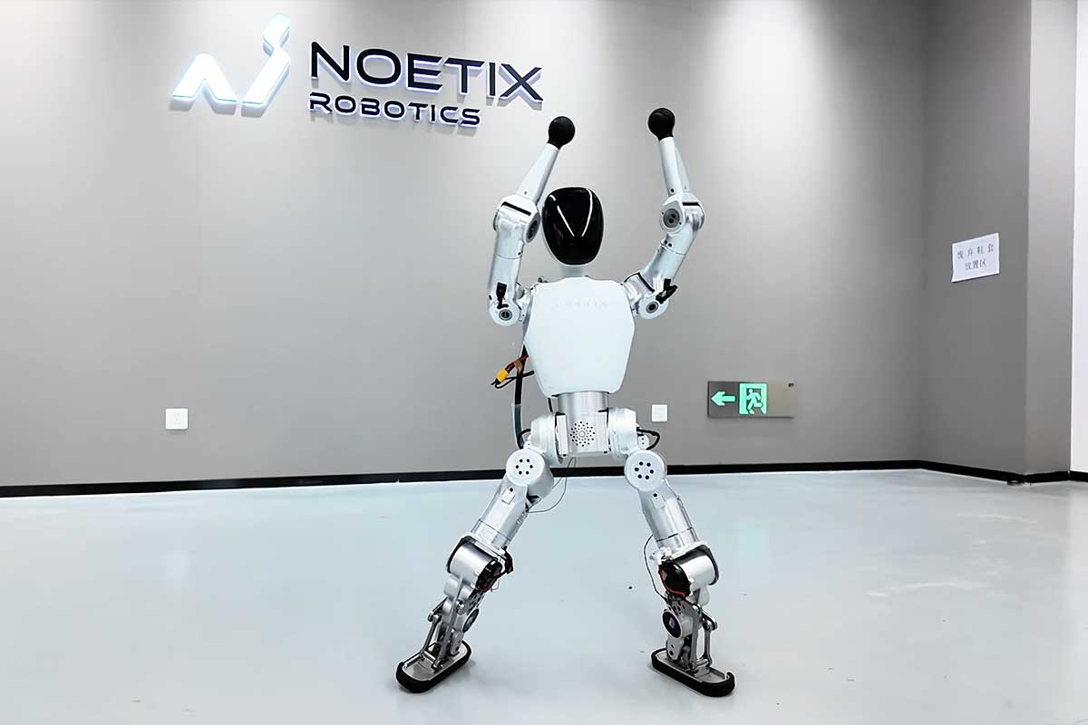 Noetix Bumi humanoid robot photographed from behind at Noetix Robotics headquarters in Beijing with company logo on gray wall, robot standing with both arms raised overhead demonstrating shoulder joint articulation range, exposed mechanical construction showing silver aluminum actuators at hip knee and ankle joints, black spherical hand end-effectors, central torso battery compartment with ventilation ports visible, white composite body panels with modular design for 12kg total weight
