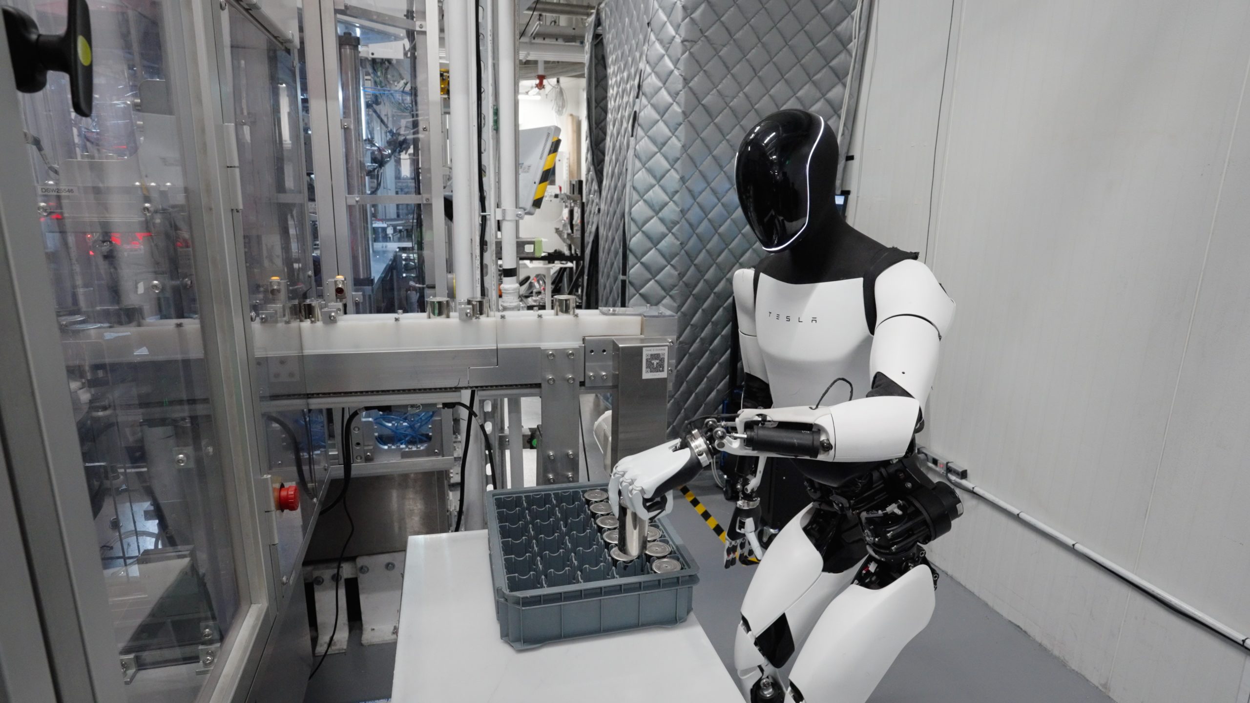 Tesla Optimus humanoid robot working at an industrial factory workstation, handling battery components alongside automated manufacturing equipment at Tesla Gigafactory