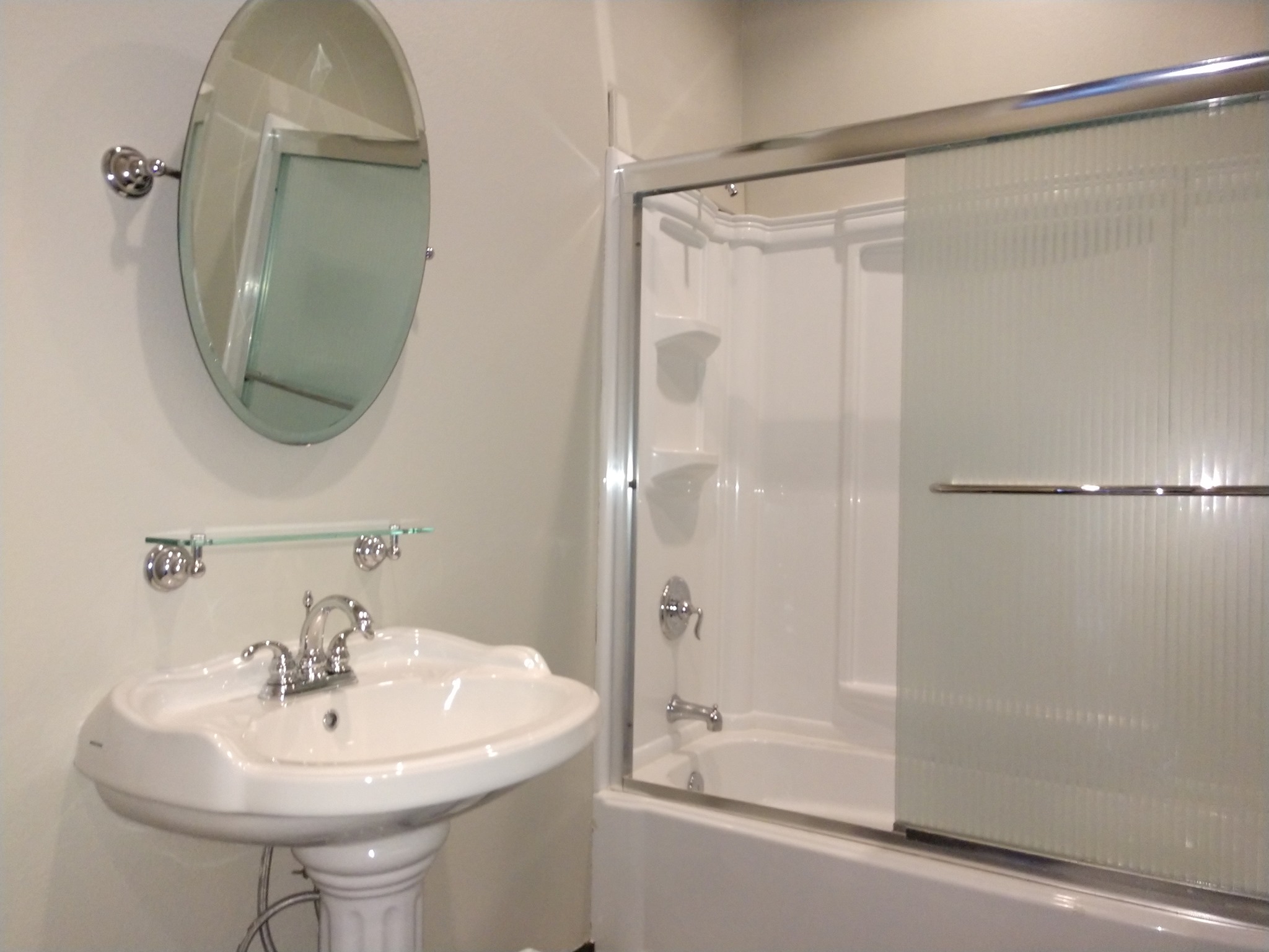 Bathroom with a white pedestal sink, oval mirror, and bathtub with sliding frosted glass door.