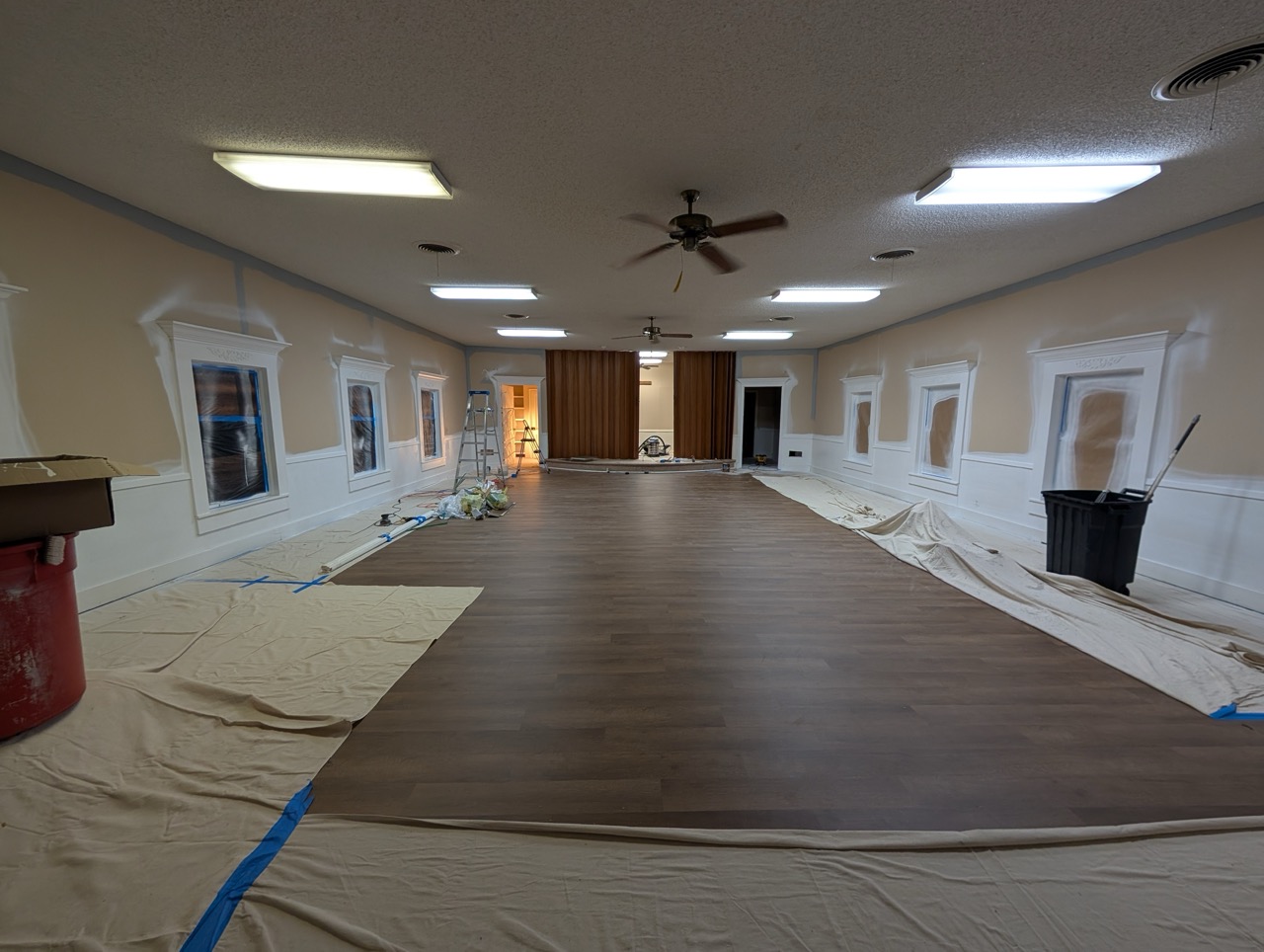 Large room under renovation with wood floor, covered windows, drop cloths, ladders, and ceiling fans.
