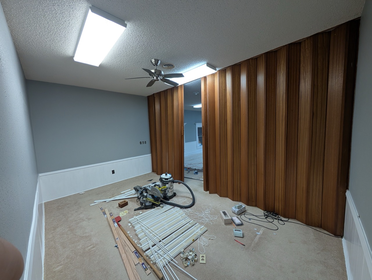Room under renovation with carpet, fluorescent lights, ceiling fan, wood panel wall, and scattered tools and materials.