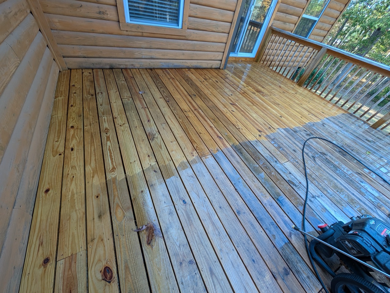 Partially cleaned wooden deck with a pressure washer on the floor near a log cabin wall and railing.